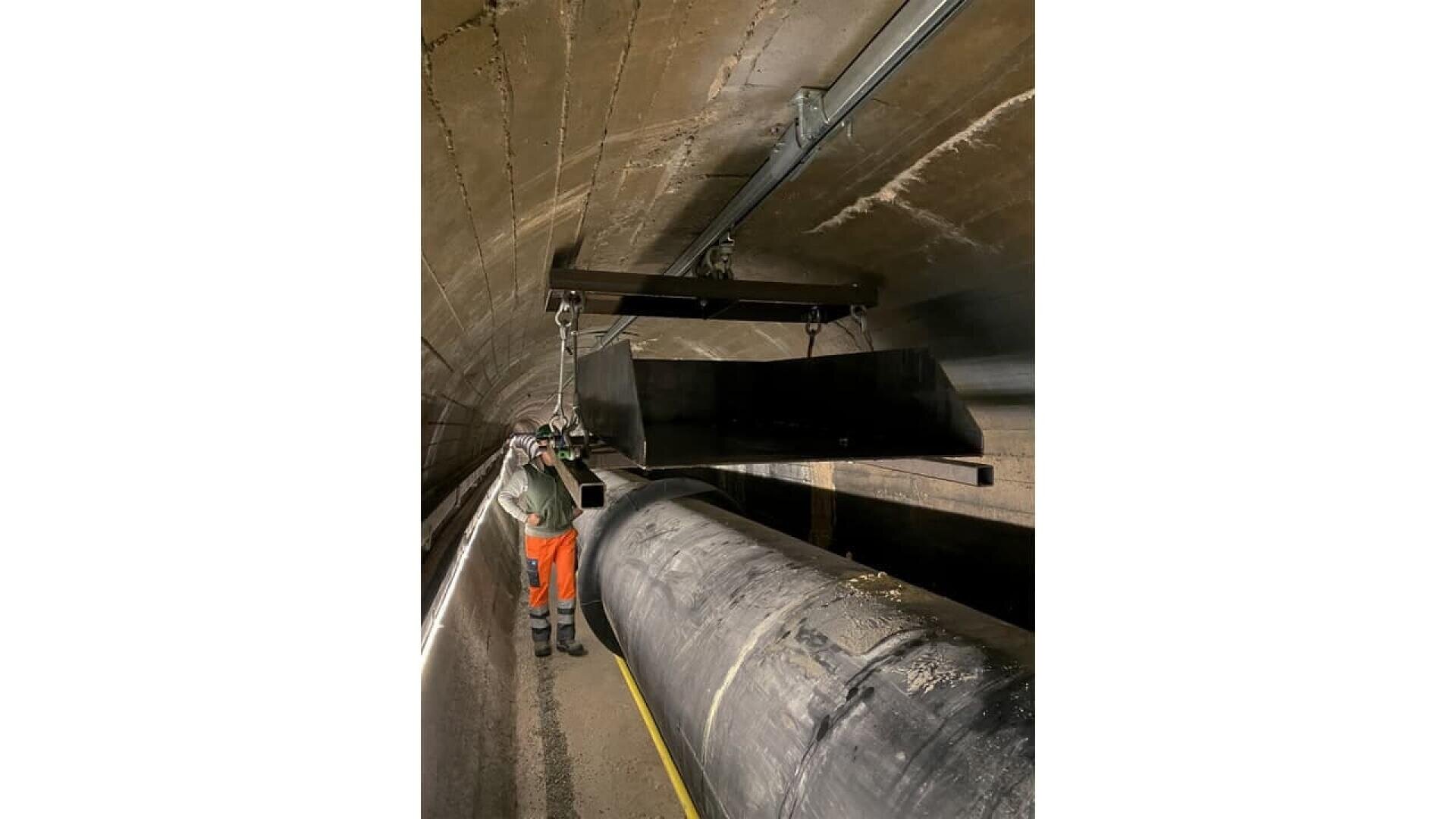 A worker wearing orange pants, a reflective vest, and a helmet stands in a tunnel next to a large pipe and beneath a suspended black metal chute attached to the ceiling.