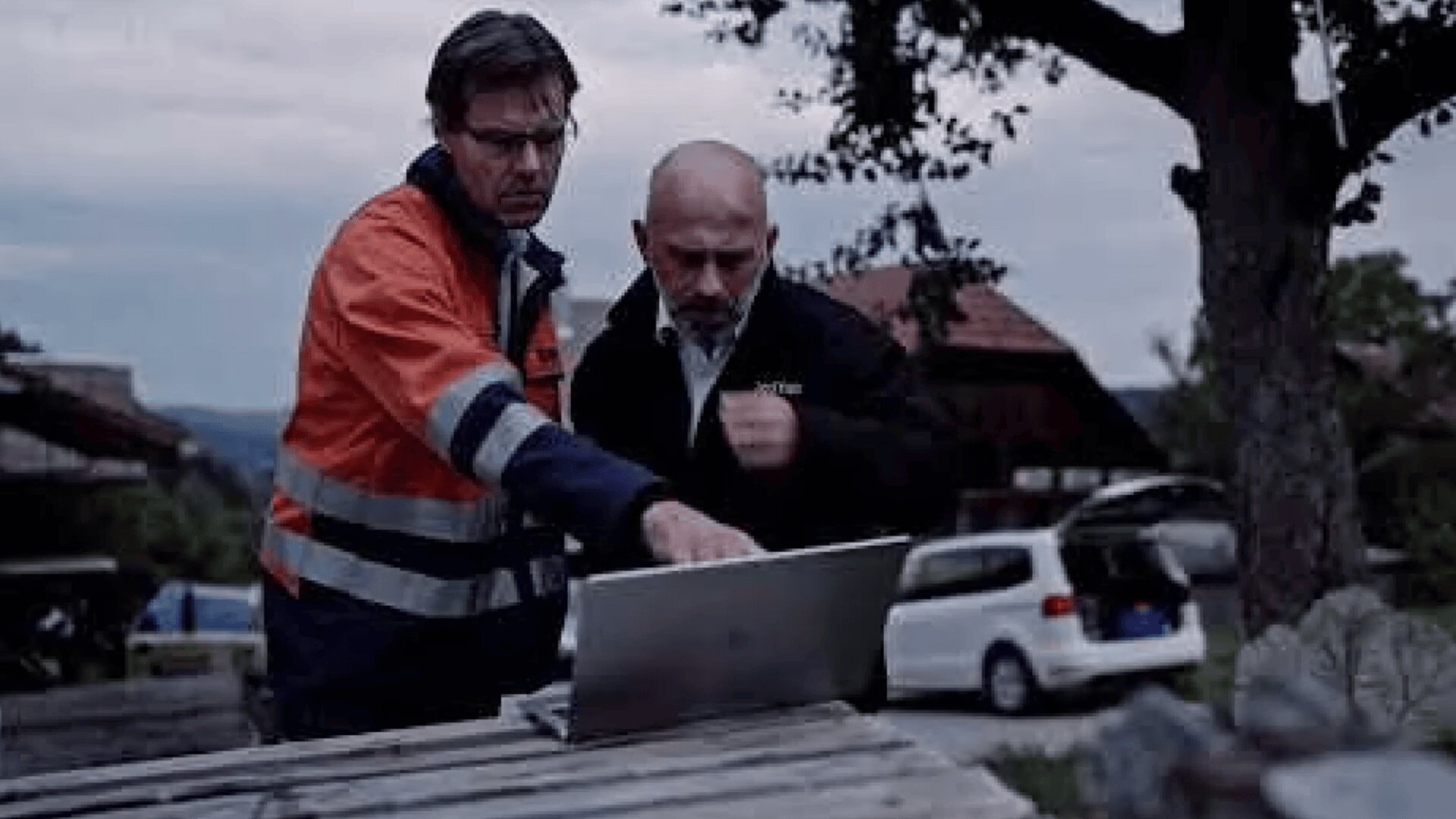 Two men stand outdoors by a wooden table, looking intently at a laptop. One, wearing an orange safety jacket, points at the screen while the other, in dark clothing, watches. A white car and trees are visible in the background.
