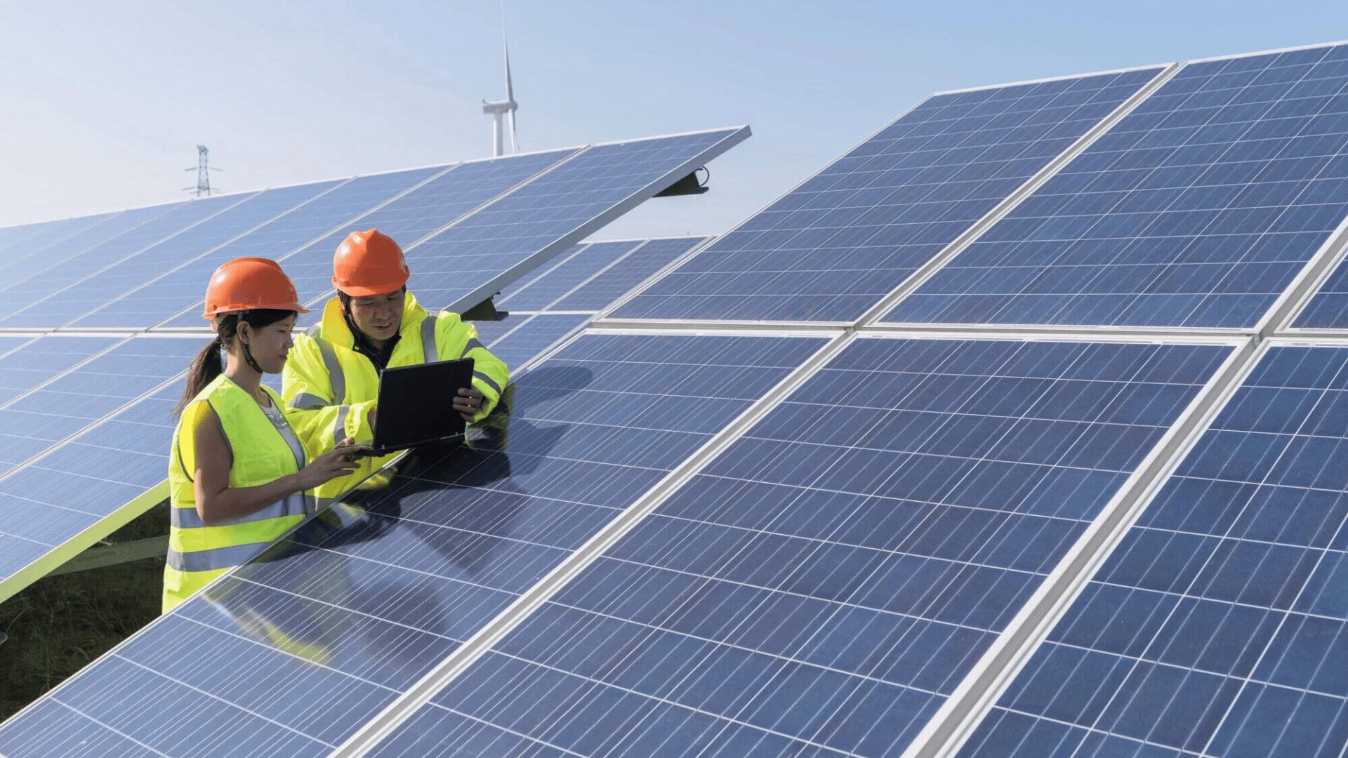 Zwei Arbeiter in Sicherheitswesten und orangefarbenen Helmen untersuchen an einem sonnigen Tag im Freien einen Laptop neben großen Solarzellen, während im Hintergrund Windräder zu sehen sind.