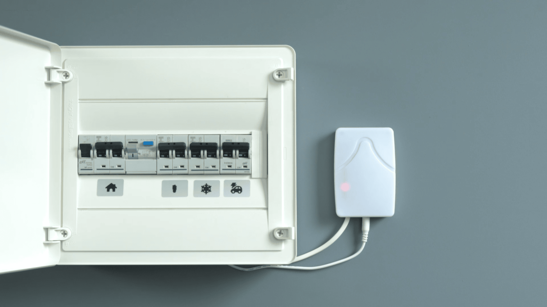 An electrical circuit breaker panel with its door open is mounted on a gray wall. Next to it is a white device with a glowing red indicator light, connected by two cables.