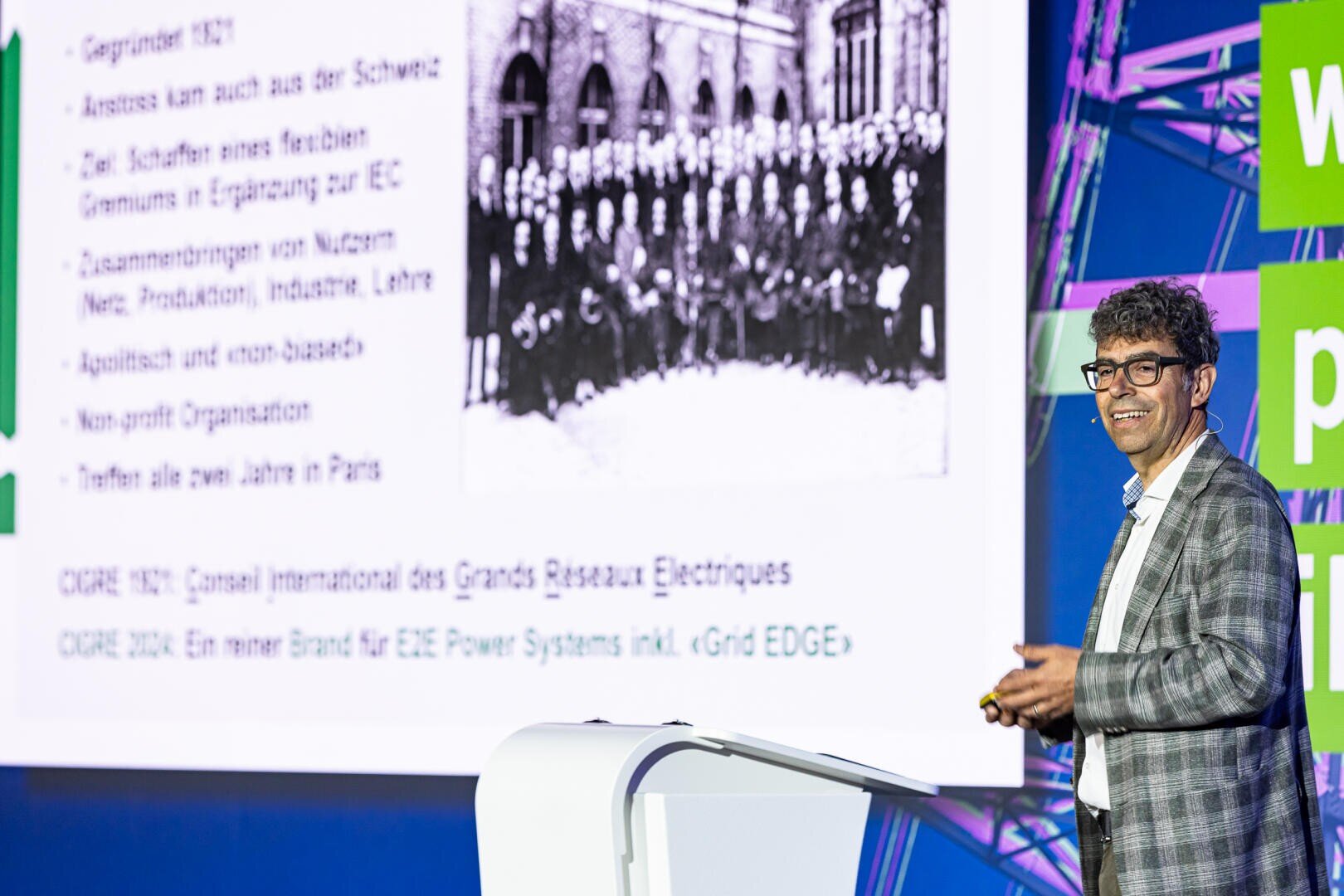Ein Mann im Anzug und mit Brille steht lächelnd an einem Podium und hält einen Vortrag vor dem Publikum. Hinter ihm ist ein großes Dia mit deutschem Text und einem historischen Gruppenfoto in Schwarz-Weiß zu sehen. Helle Bühnenlichter und grüne Akzente sind zu sehen.