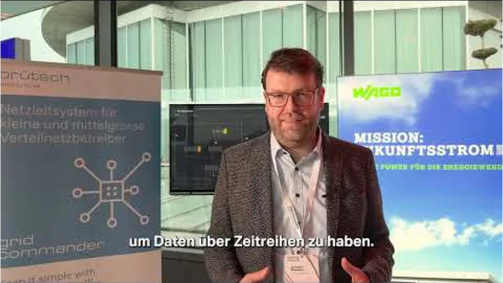 A man wearing glasses and a suit stands indoors between two banners, speaking to the camera. German subtitles appear at the bottom. Large windows and modern architecture are visible in the background.