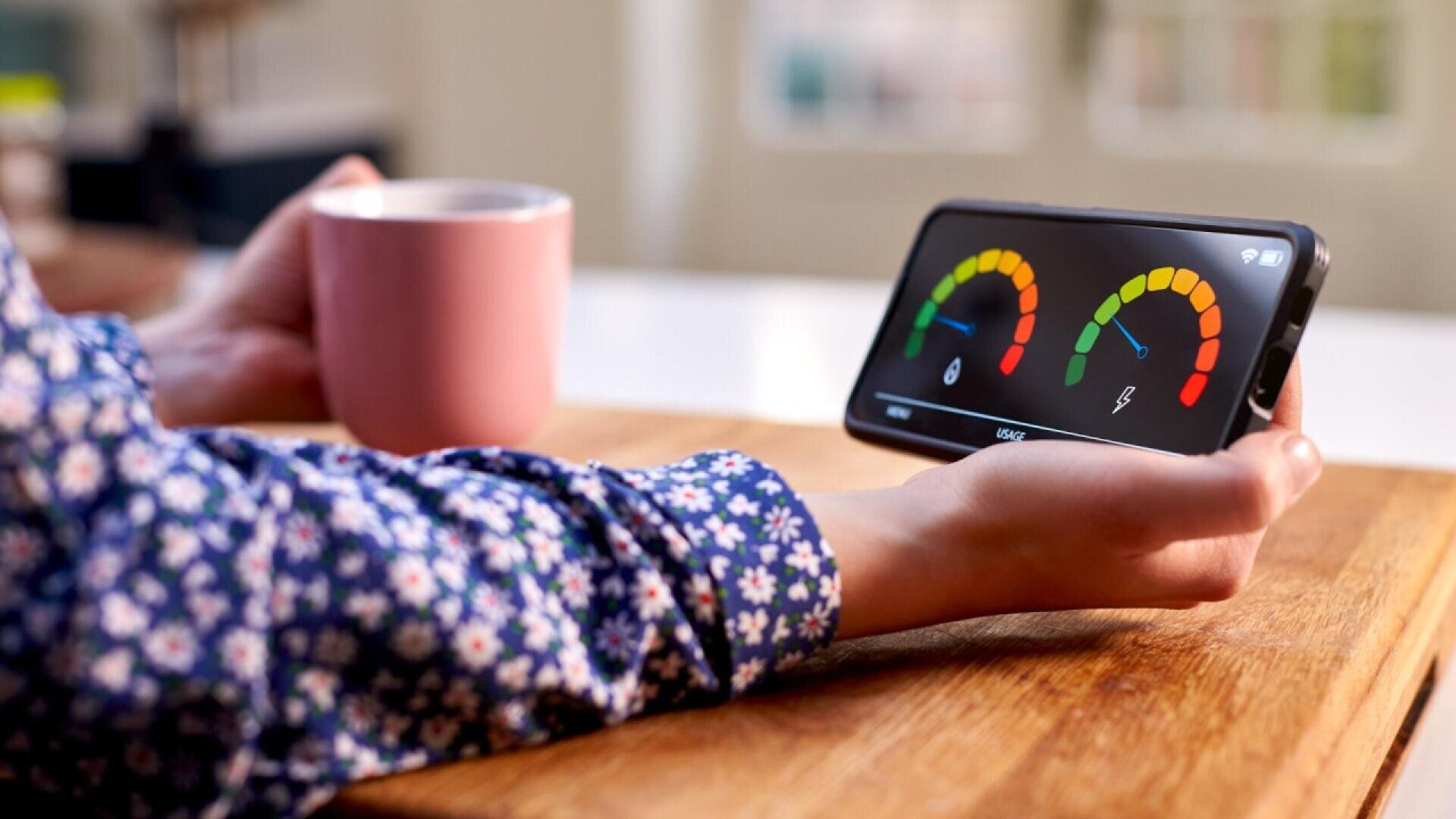 A person in a floral shirt holds a pink mug while looking at a smartphone displaying colorful dials or meters on a wooden table. The scene is indoors with a soft background.