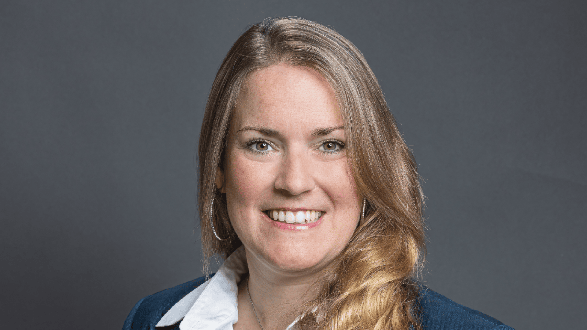 Eine Frau mit langen blonden Haaren, die einen dunklen Blazer und ein weißes Hemd mit Kragen trägt, lächelt in die Kamera vor einem schlichten dunkelgrauen Hintergrund.