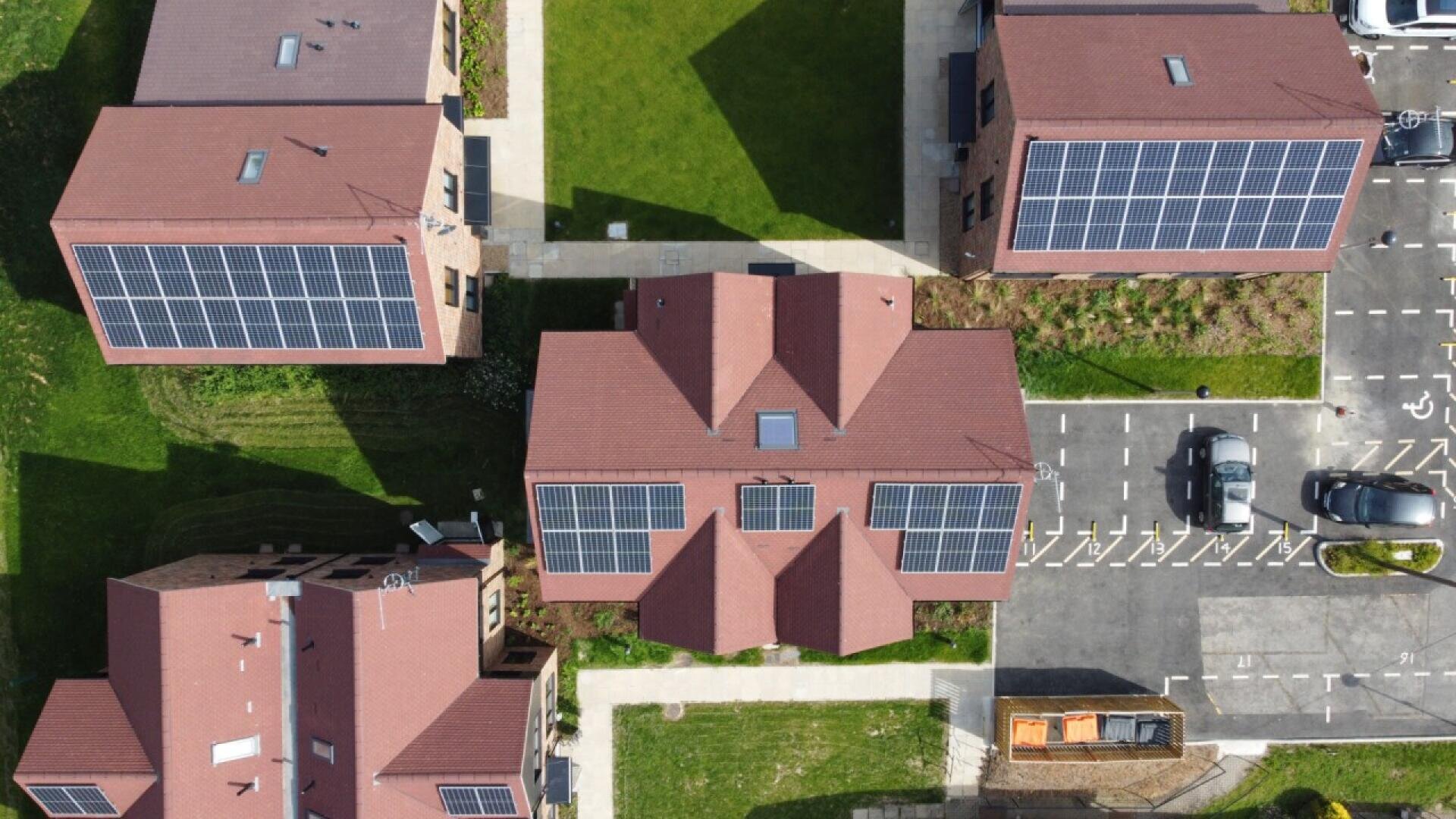 Aerial view of four buildings with brown roofs and solar panels, surrounded by green lawns and parking areas with cars and a truck. Pathways run between the buildings.
