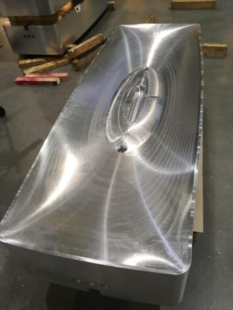 Eine große, polierte Metallform mit einer glatten, ovalen Vertiefung in der Mitte ruht auf einem Betonboden, der von Holzplanken umgeben ist. Die Oberfläche weist sichtbare Bearbeitungsspuren und eine spiegelnde Oberfläche auf.