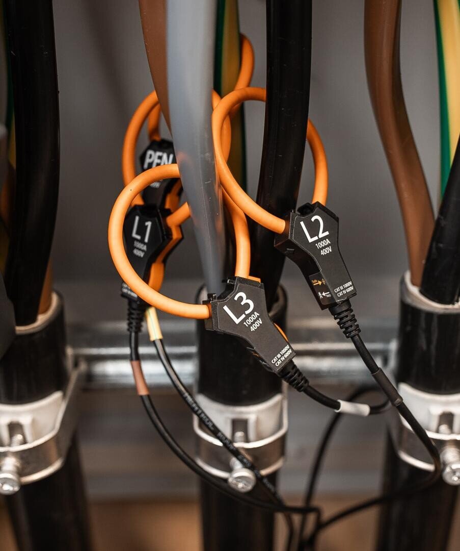 Nahaufnahme von elektrischen Kabeln mit orangefarbenen Schleifen und schwarzen Steckern mit den Bezeichnungen L1, L2 und L3, die an dicken schwarzen Drähten in einer industriellen oder technischen Umgebung befestigt sind.