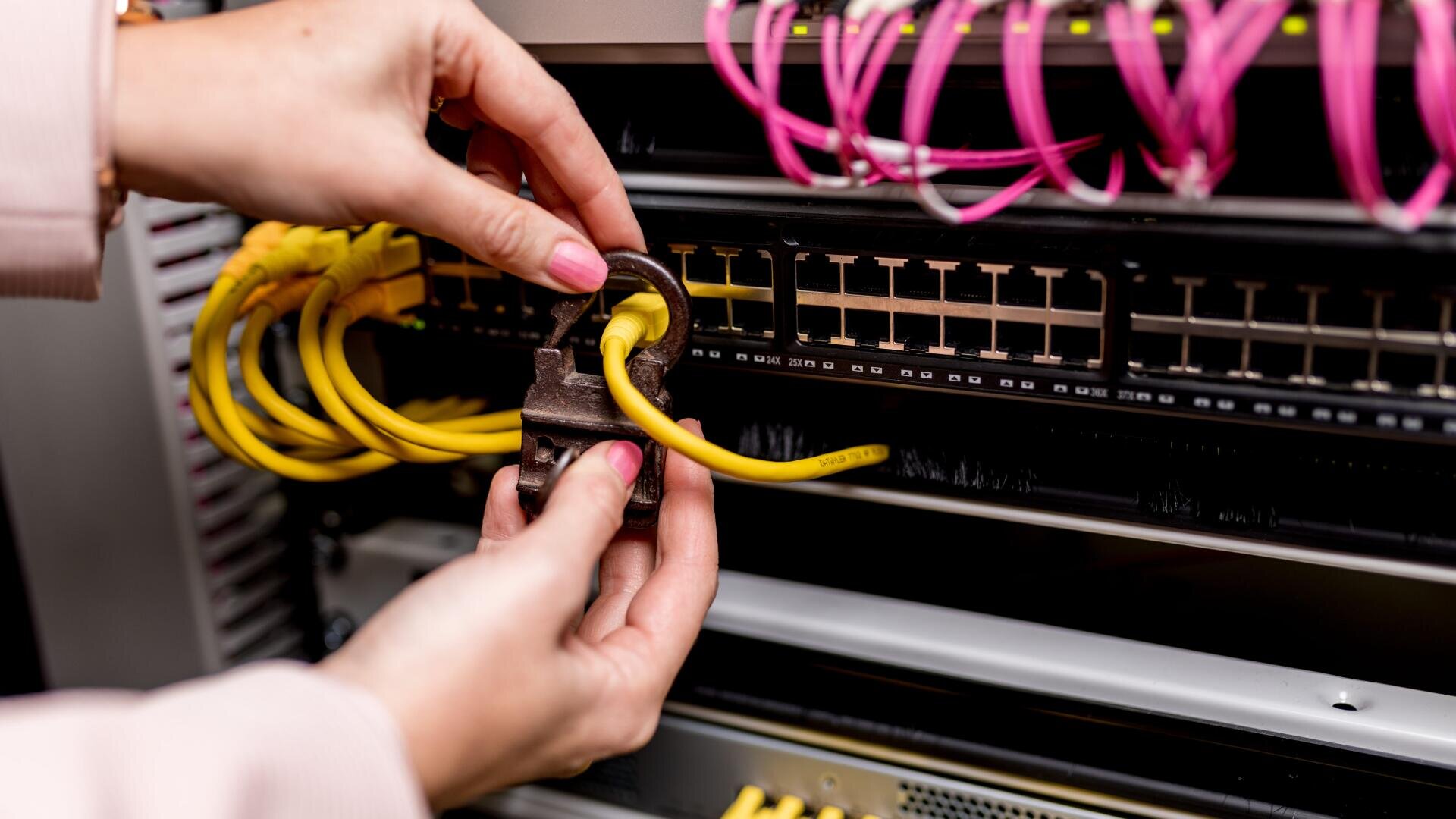 Eine Person bringt ein Vorhängeschloss an einem gelben Netzwerkkabel an, das mit einem Serverschrank verbunden ist, und symbolisiert damit Datensicherheit oder Netzwerkschutz. Über dem verschlossenen Kabel sind auch rosa Kabel zu sehen.