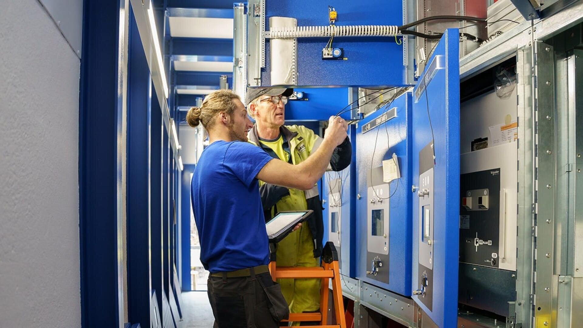 Zwei Techniker arbeiten zusammen, um elektrische Schalttafeln in einer Industrieanlage zu überprüfen und einzustellen. Der eine hält ein Tablet in der Hand, während der andere auf einer Leiter steht und mit Kabeln an den blauen Schalttafeln arbeitet.