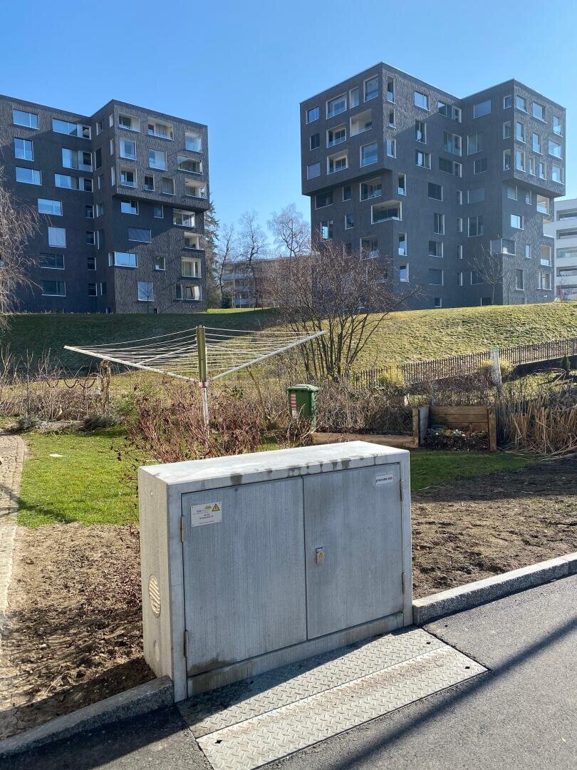Ein Stromkasten aus Beton steht in der Nähe des Bürgersteigs vor einem grasbewachsenen Abhang. Im Hintergrund stehen moderne, dunkle Wohnhäuser unter einem klaren blauen Himmel.