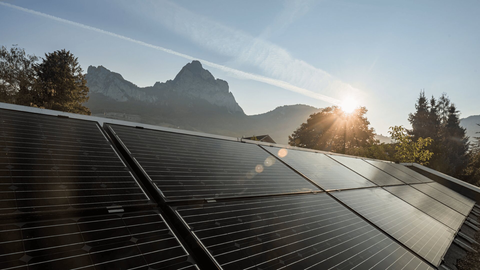 Solarmodule auf einem Dach, die Sonne geht hinter Bäumen und einer Bergkette im Hintergrund auf, unter einem klaren blauen Himmel.