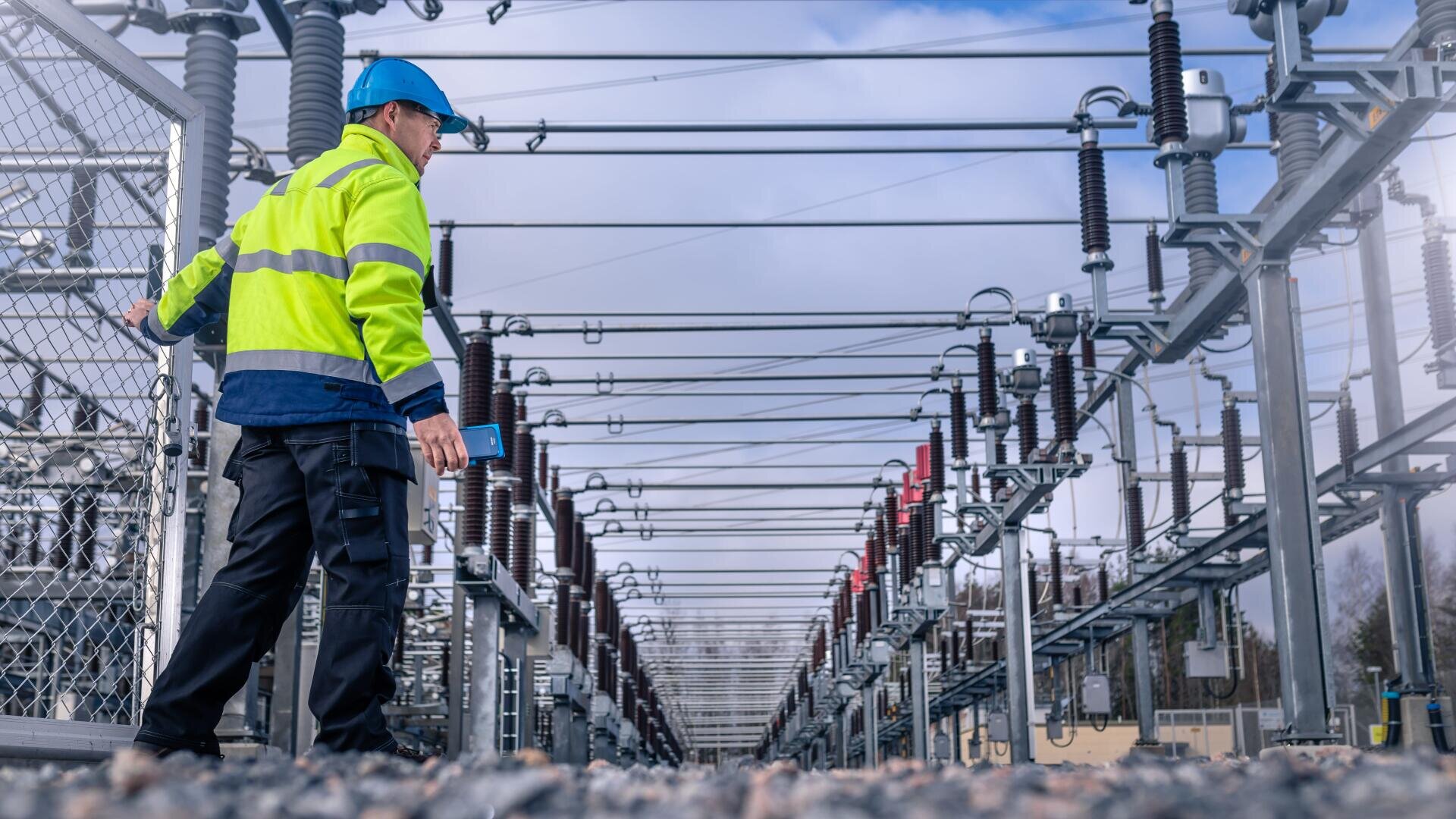 A worker in a high-visibility jacket and blue helmet inspects equipment at an outdoor electrical substation filled with power lines, insulators, and metal structures under a cloudy sky.