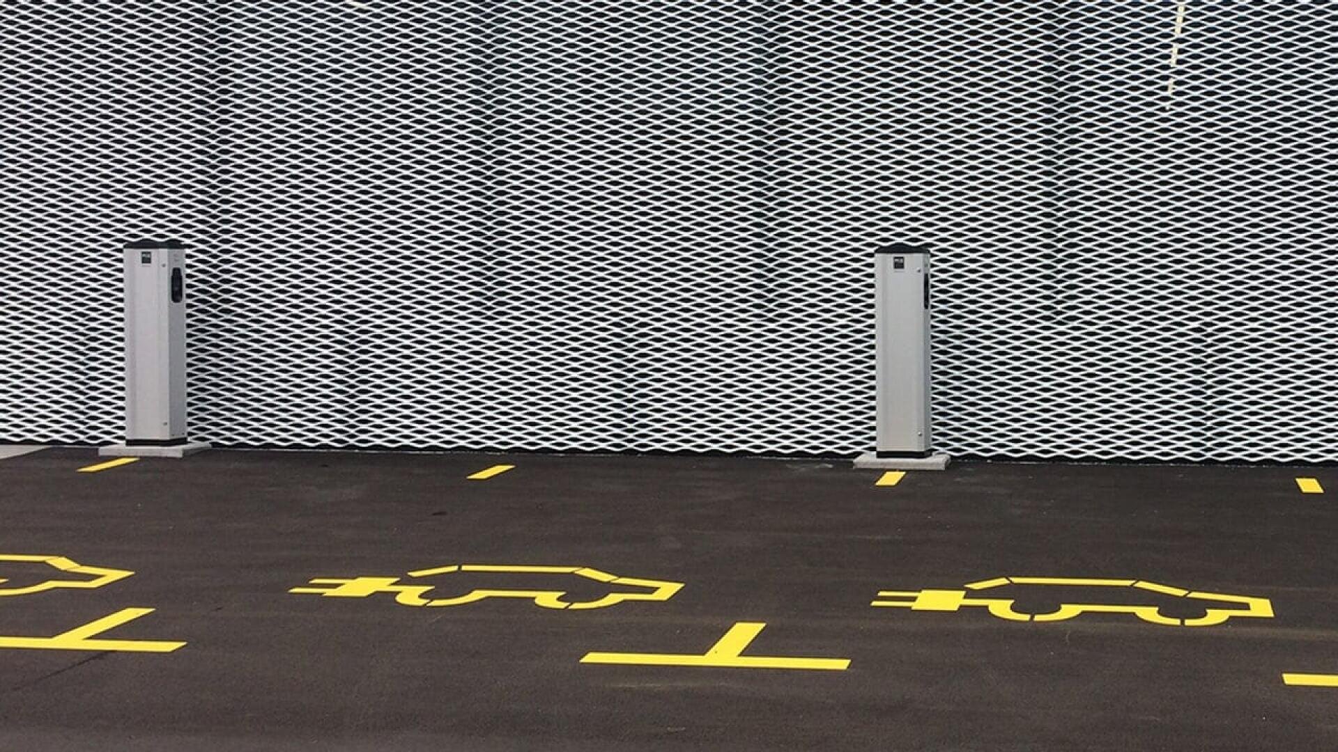 Two electric vehicle charging stations stand in front of a metal mesh wall, with yellow painted outlines of cars and charging symbols on the asphalt indicating designated EV parking spaces.