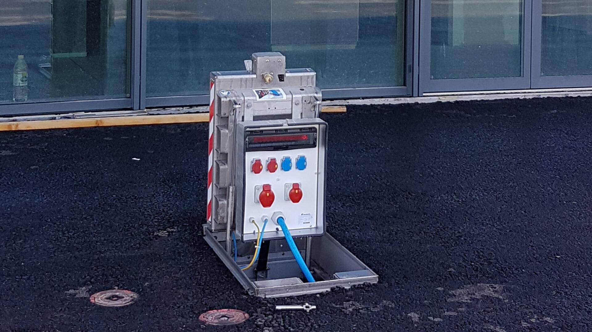 Ein aufklappbarer Außenkasten mit mehreren Steckdosen und Steckern ist auf einer gepflasterten Fläche vor einem Gebäude mit großen Glasfenstern installiert. Der Kasten ist teilweise geöffnet und gibt den Blick auf farbige Steckdosen und Kabel frei.
