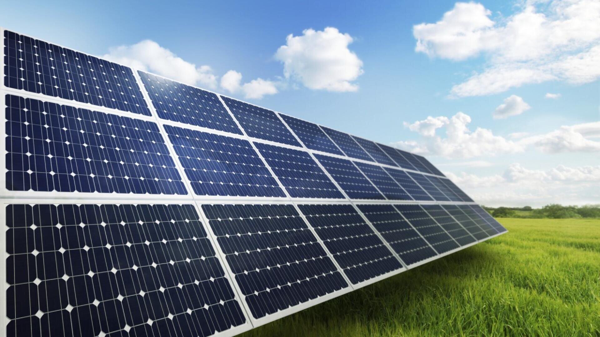 A row of solar panels stands in a green grassy field under a blue sky with scattered white clouds, capturing sunlight for renewable energy.
