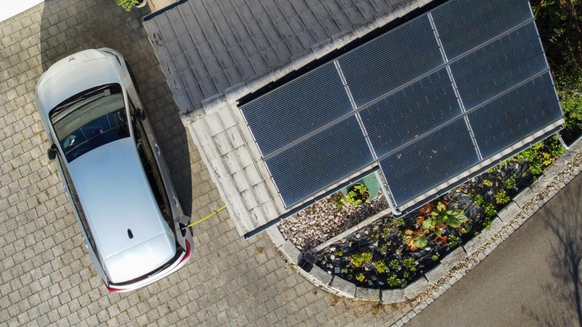 Aerial view of a silver car parked next to a building with a rooftop covered in solar panels, connected by a charging cable; nearby, there is a landscaped garden with plants and a paved driveway.
