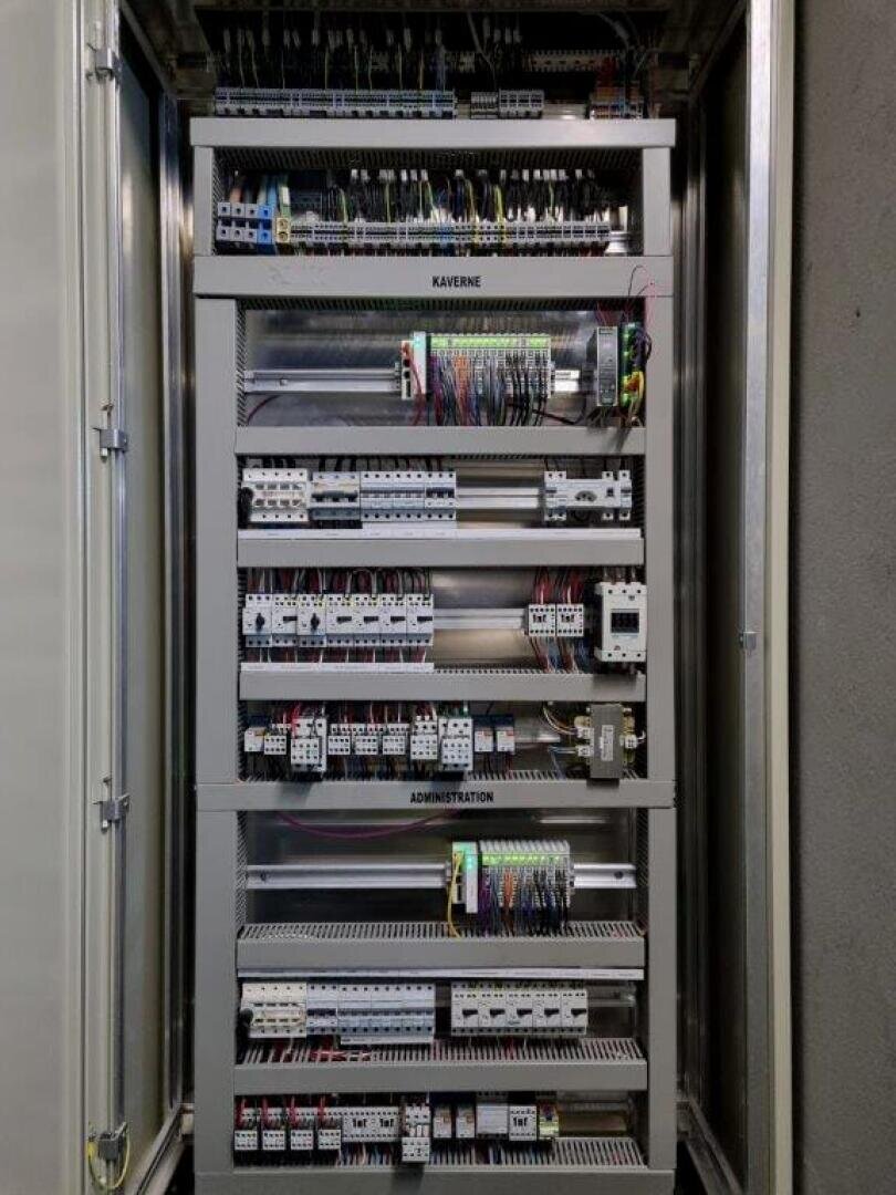 Eine offene elektrische Schalttafel mit ordentlich angeordneten Drähten, Leistungsschaltern, Relais und beschrifteten Bauteilen, darunter Abschnitte mit der Aufschrift KAVERNE und ADMINISTRATION.
