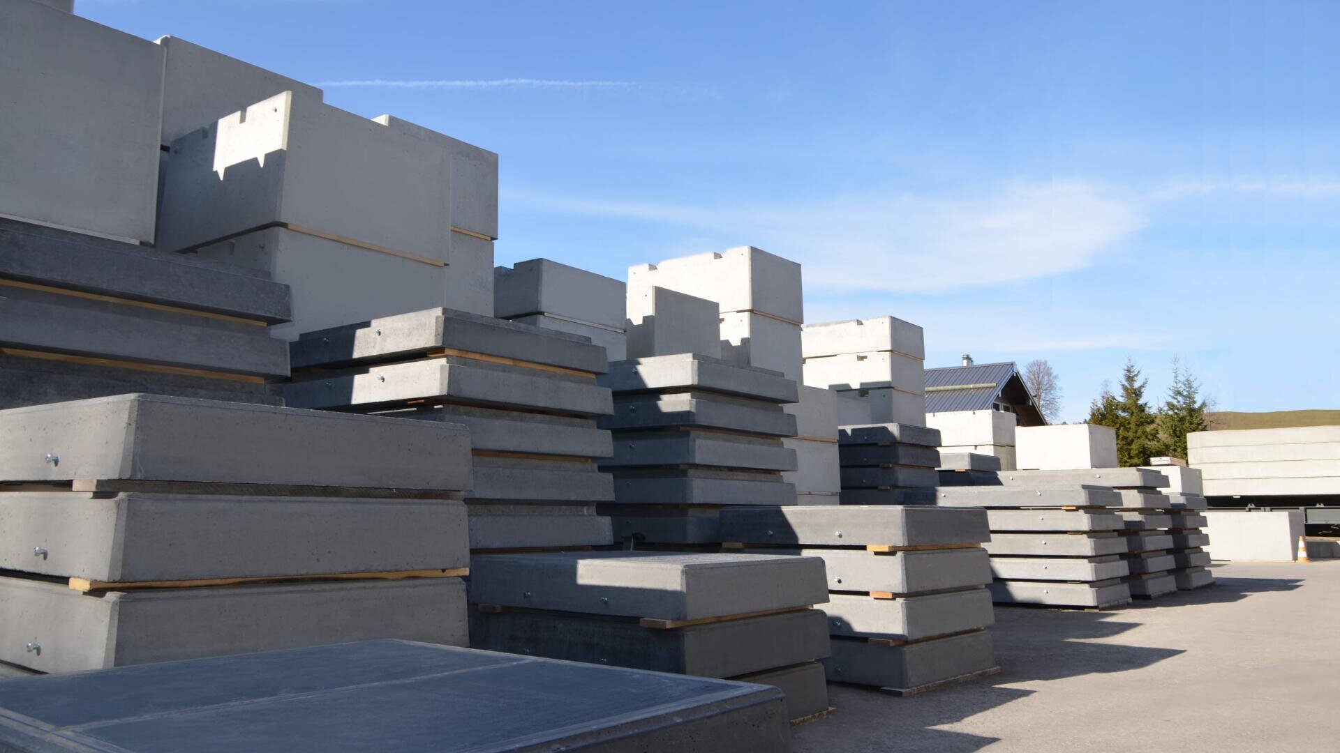 Stapel von großen rechteckigen Betonblöcken sind auf einem Lagerplatz im Freien unter einem klaren blauen Himmel angeordnet. Im Hintergrund sind einige Bäume und Gebäude zu sehen.