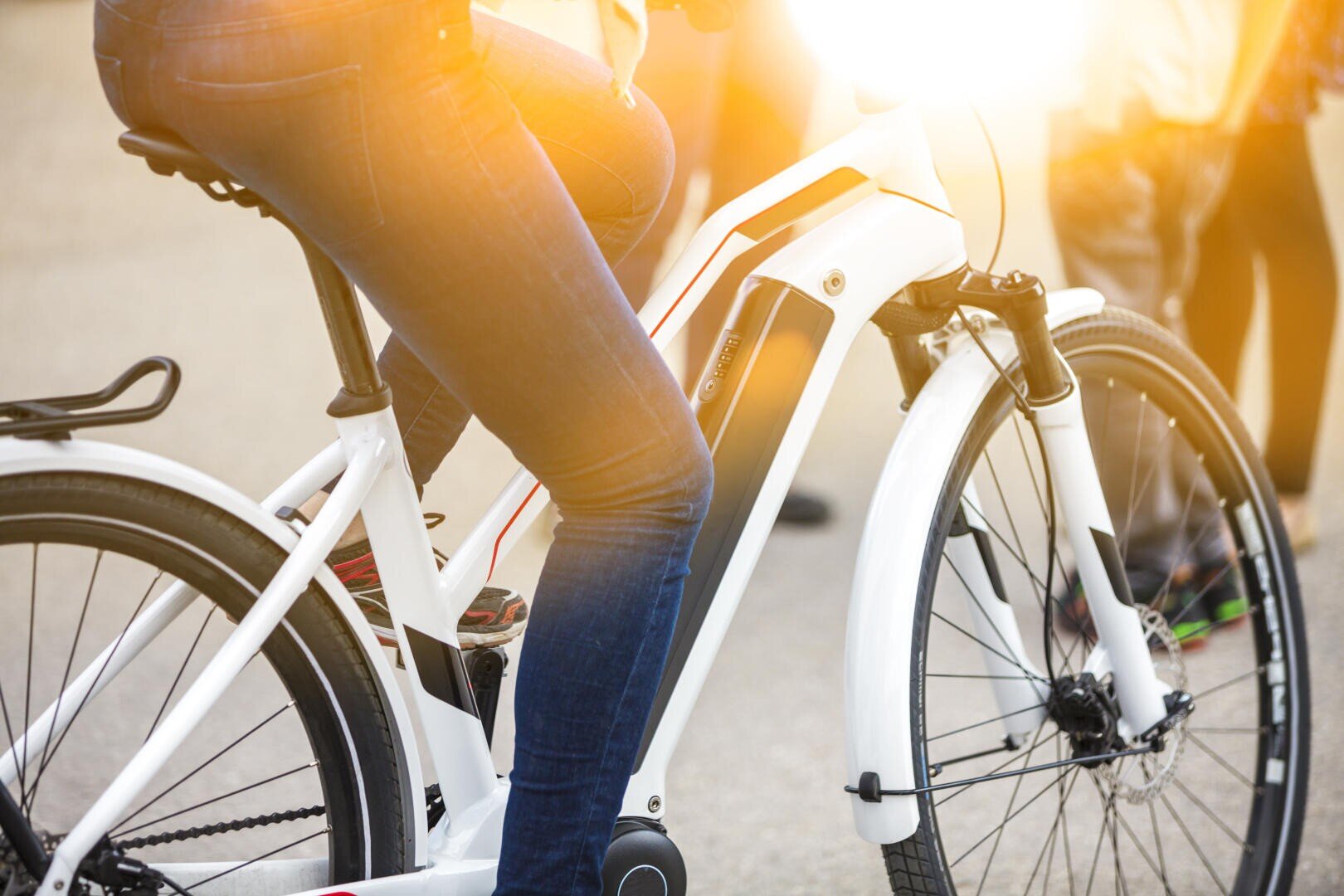A person wearing jeans is sitting outside on a white e-bike; other people can be seen blurred in the background, and sunlight is shining on the scene.