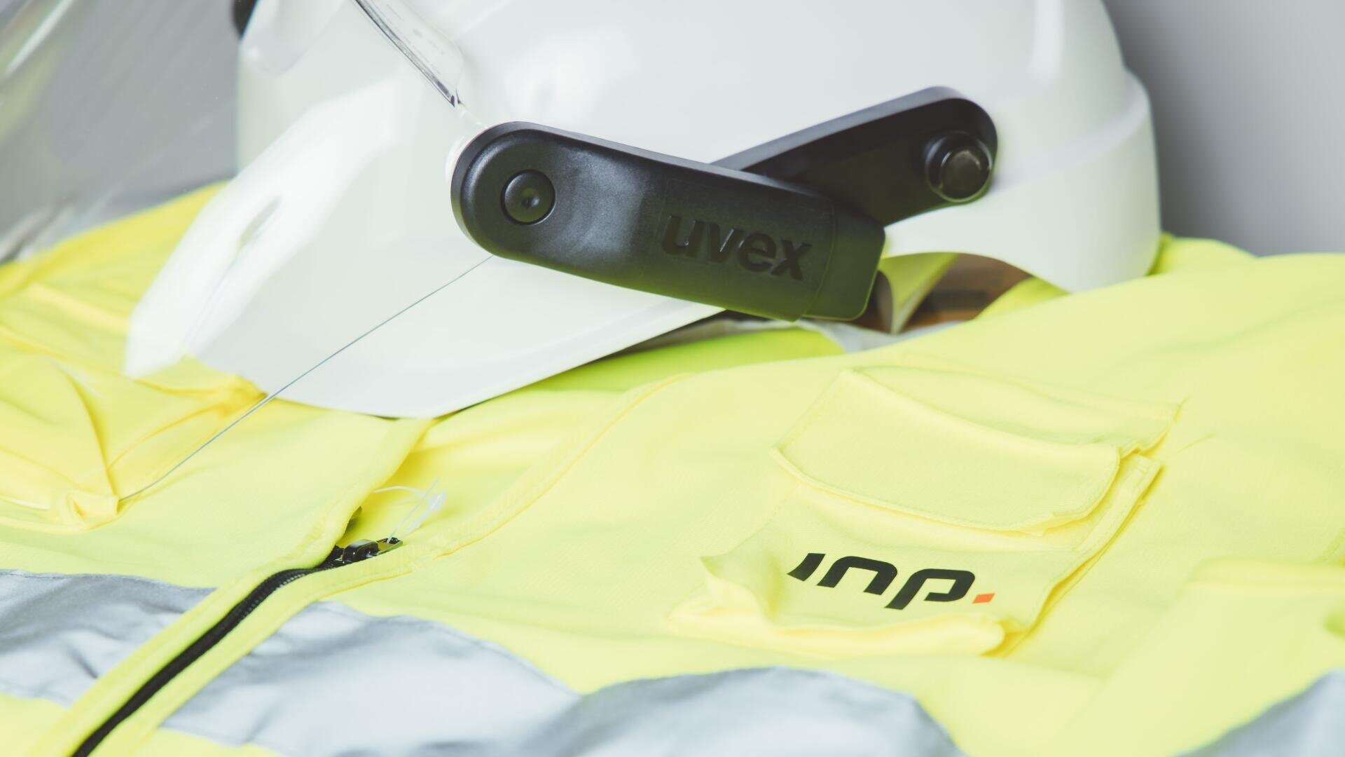 Nahaufnahme eines weißen Schutzhelms mit Visier und einer auffälligen gelben Sicherheitsjacke mit reflektierenden Streifen und einer sichtbaren Tasche. Der Helm trägt das uvex Branding an der Seite.