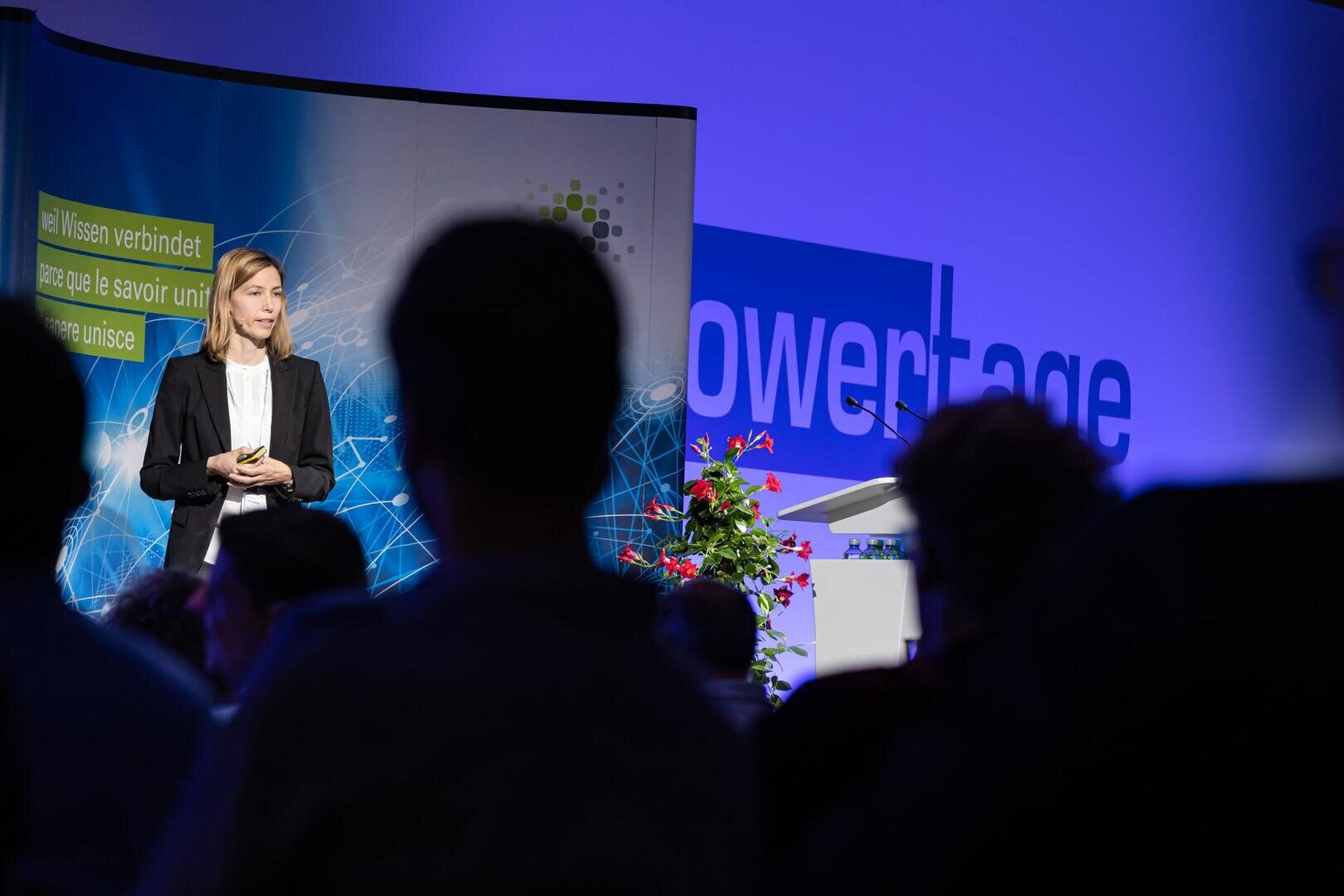 Eine Frau im Anzug steht auf der Bühne und hält einen Vortrag, in der Nähe befinden sich ein großer Bildschirm und ein Podium. Die Zuhörer sitzen im Vordergrund. Im Hintergrund sind blaue Lichter und das Wort "Powertage" zu sehen.