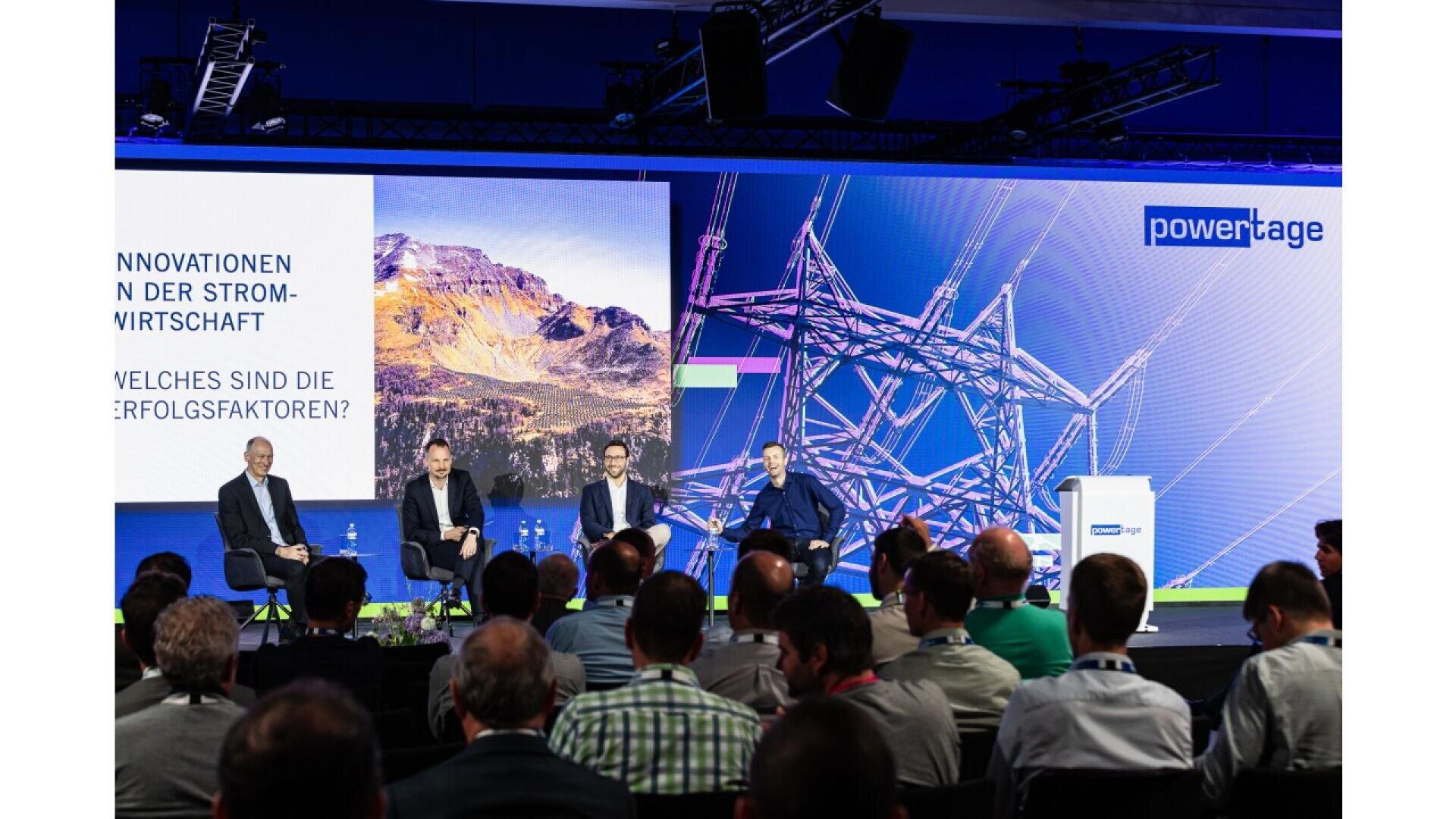 A panel of four speakers sits on stage facing an audience at a conference. Behind them are large screens displaying a mountain landscape and power lines, with “powertage” branding and German text about innovation in the energy sector.