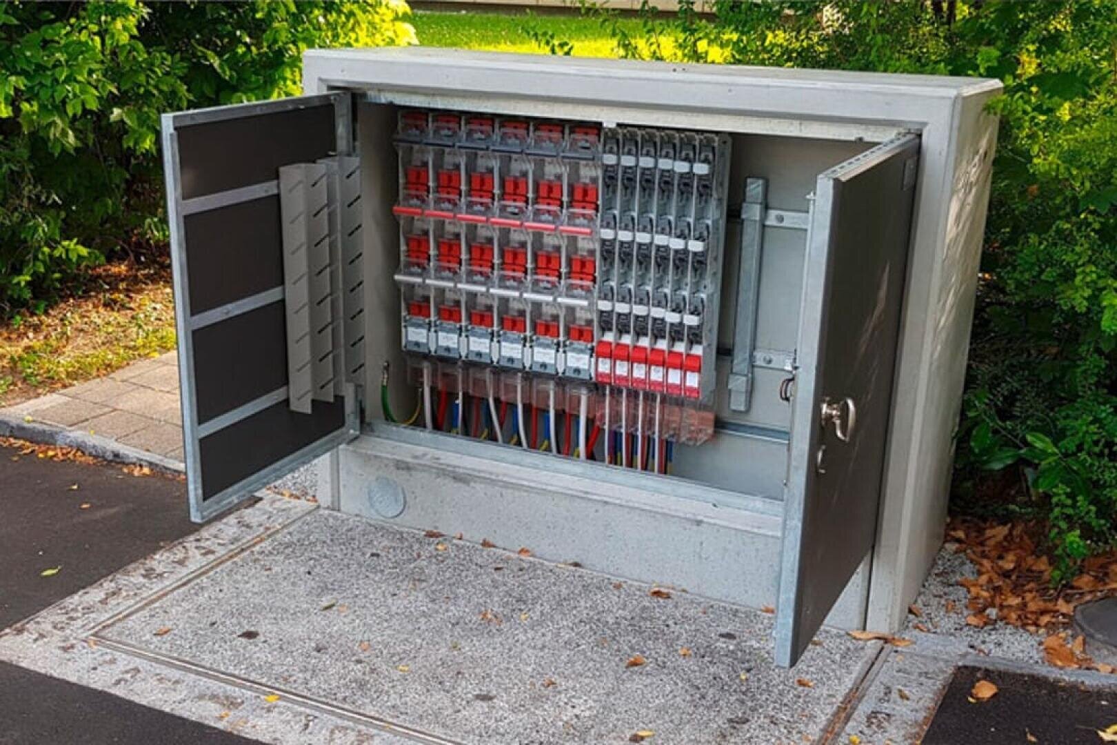 Ein offener Verteilerkasten im Freien mit mehreren Schaltern, Drähten und Leistungsschaltern auf einer Betonplatte in der Nähe einer grünen, begrünten Fläche. Die Türen des Kastens sind offen und geben den Blick auf die interne Verkabelung frei.