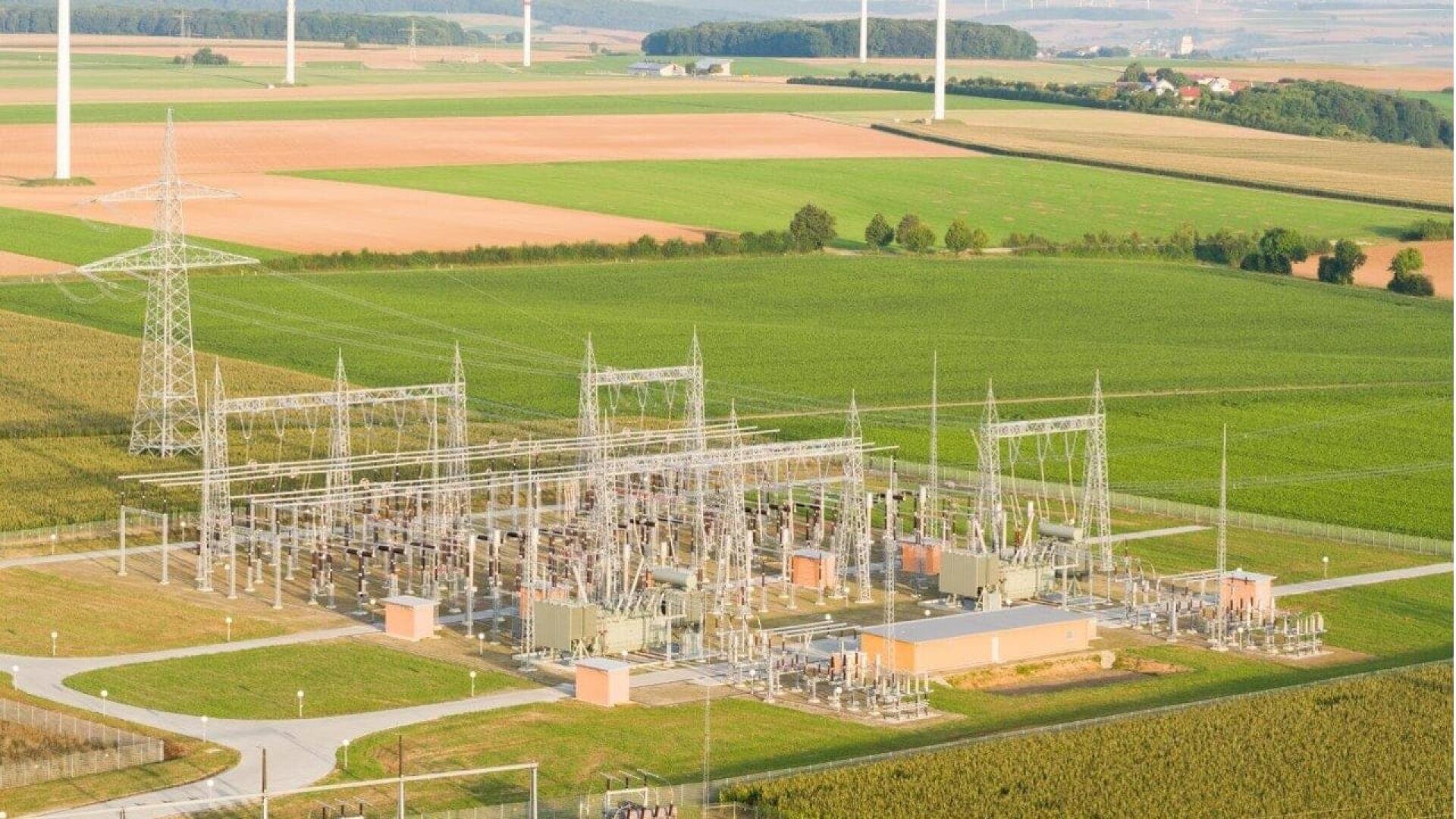 Aerial view of an electrical substation surrounded by farmland and green fields, with power lines and pylons extending into the distance. Wind turbines are visible on the horizon.