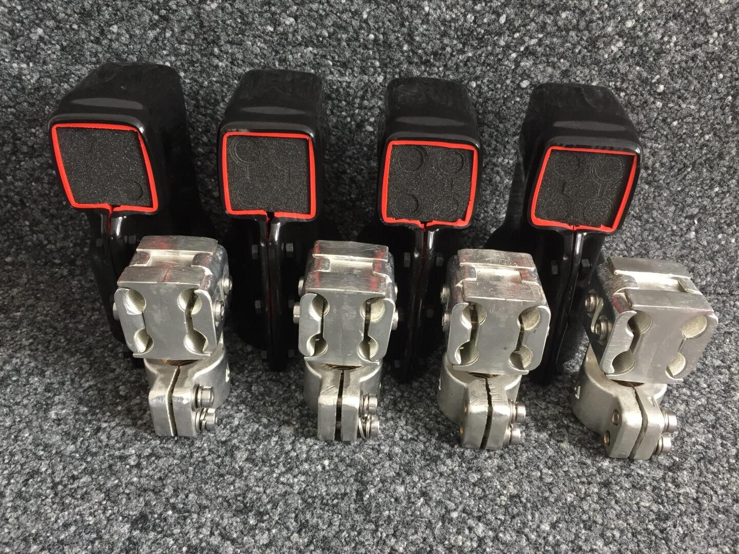 Vier schwarze Gurtschlösser mit roter Verzierung sind hinter vier metallischen Gurtschlossmechanismen auf einer grauen Teppichoberfläche aufgereiht.