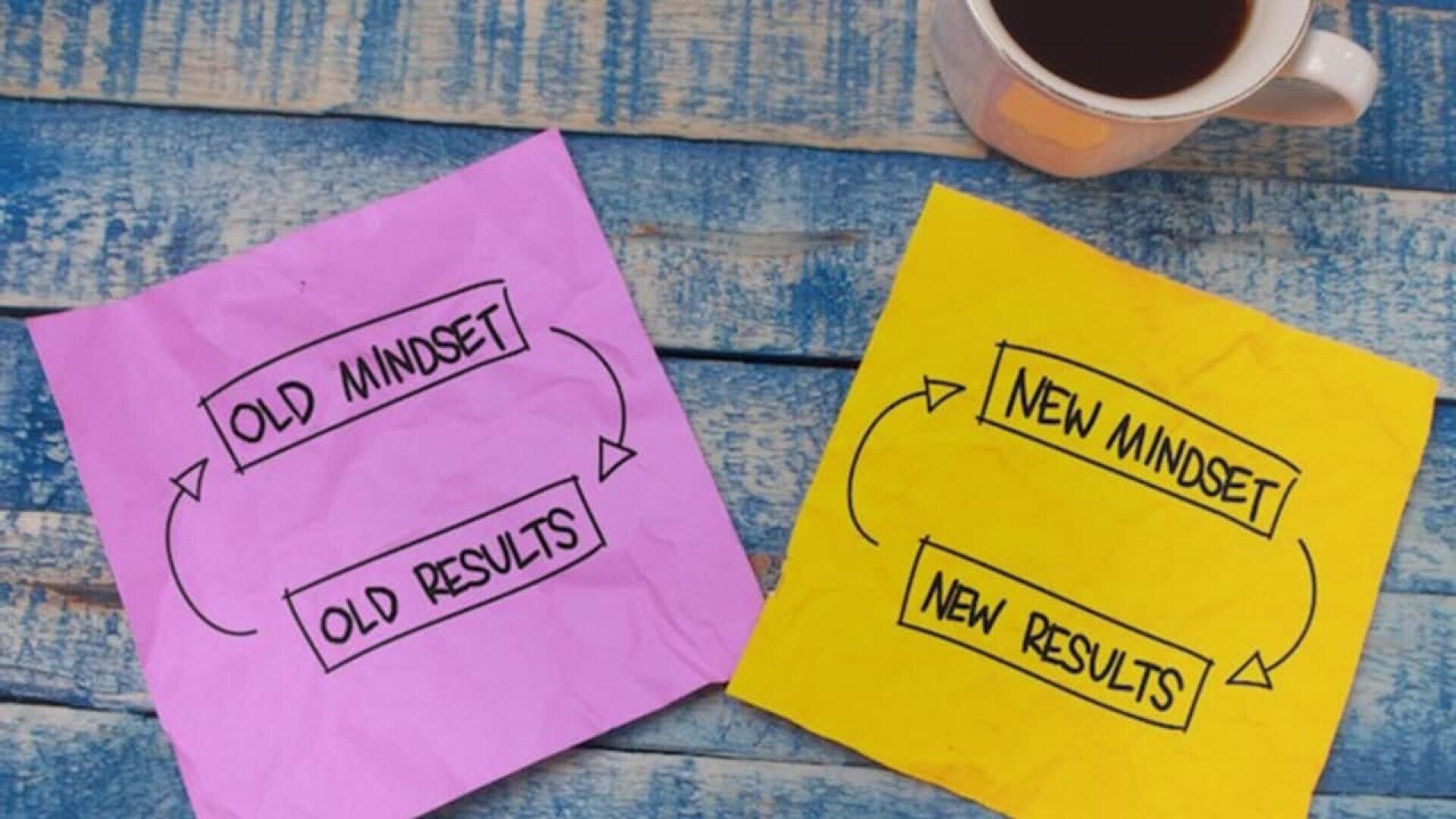 Two sticky notes on a wooden surface: one pink note reads Old mindset → Old results, and one yellow note reads New mindset → New results. A cup of coffee sits nearby.