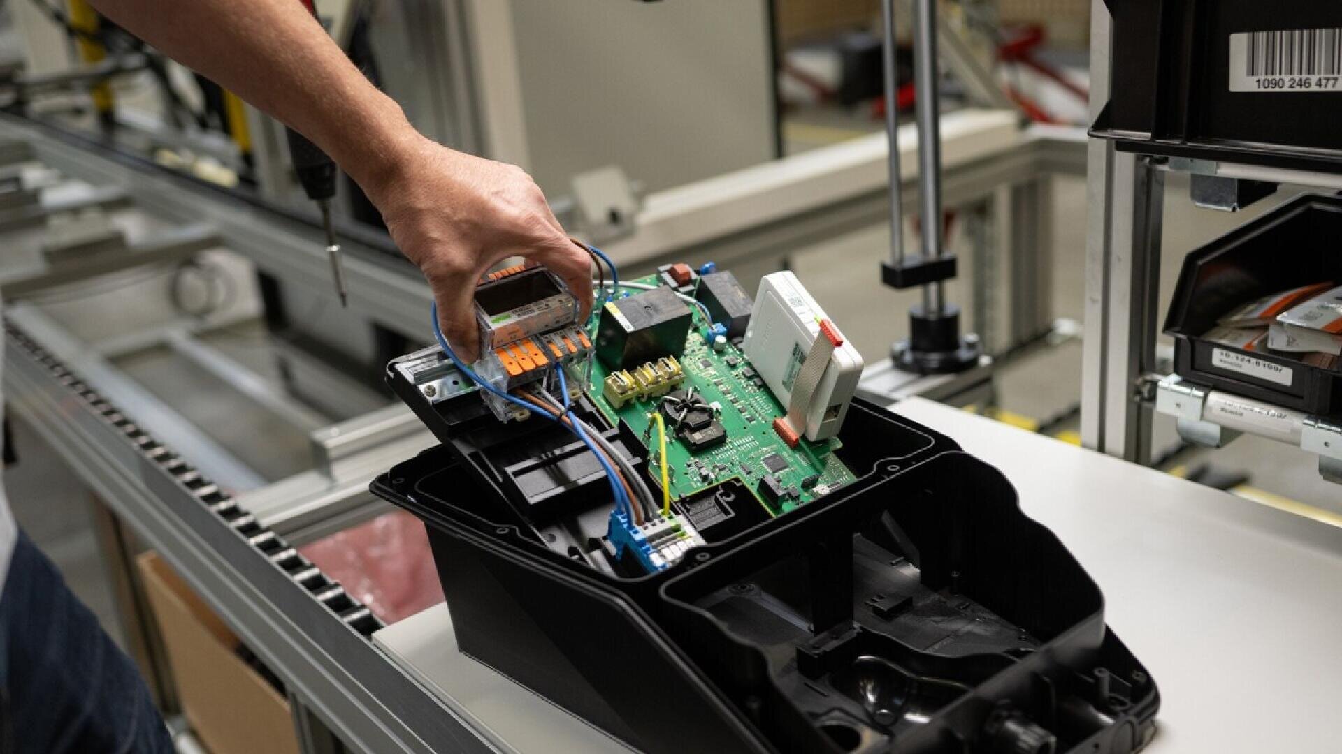 A person’s hand is assembling or inspecting an open electronic device with visible circuit boards and wires on a workbench in a factory or workshop setting.