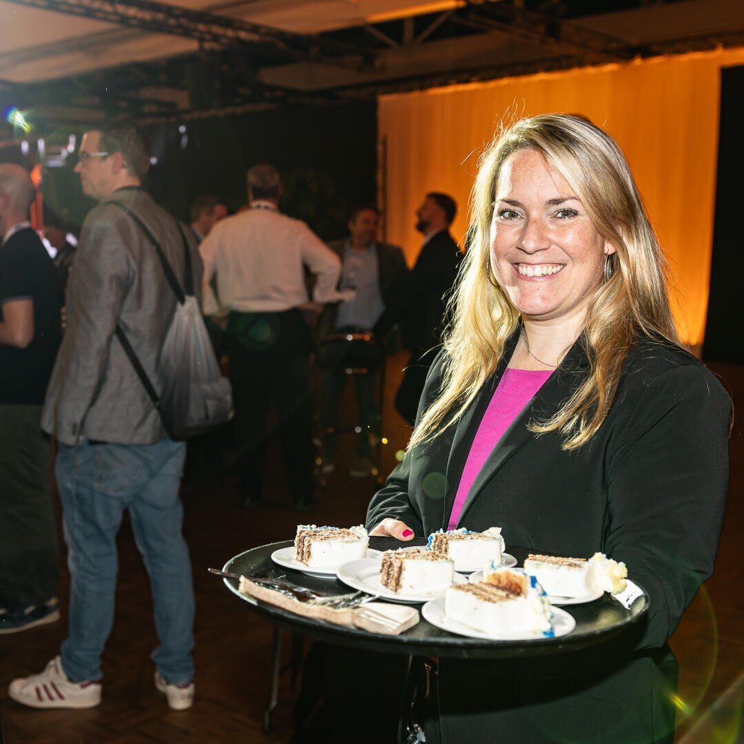 Eine lächelnde Frau in einem schwarzen Blazer hält ein Tablett mit Kuchentellern bei einer Indoor-Veranstaltung, während sich im Hintergrund bei warmer Beleuchtung Menschen treffen.