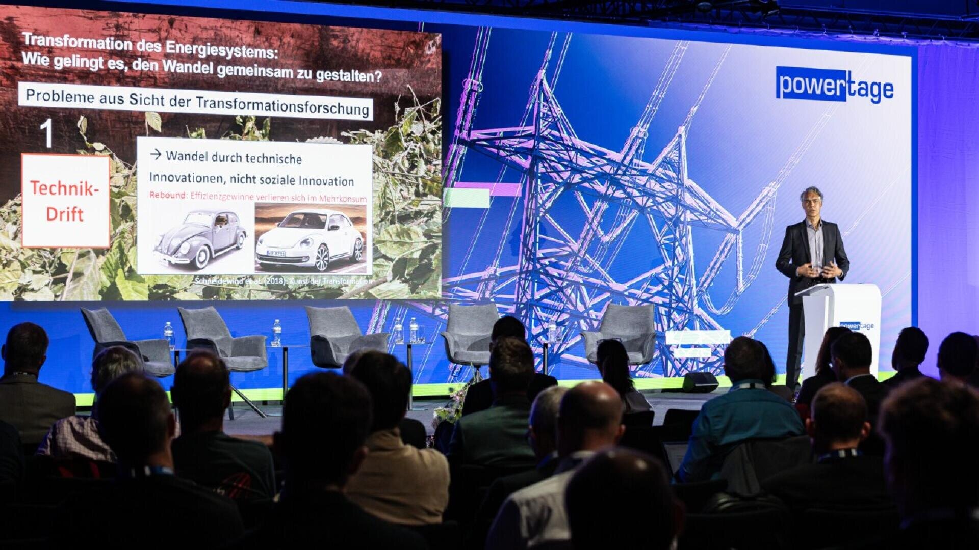 A speaker stands on stage presenting to an audience. Behind him, a large screen shows a slide with German text about energy transformation, images of cars, and an illustration of a power line structure.