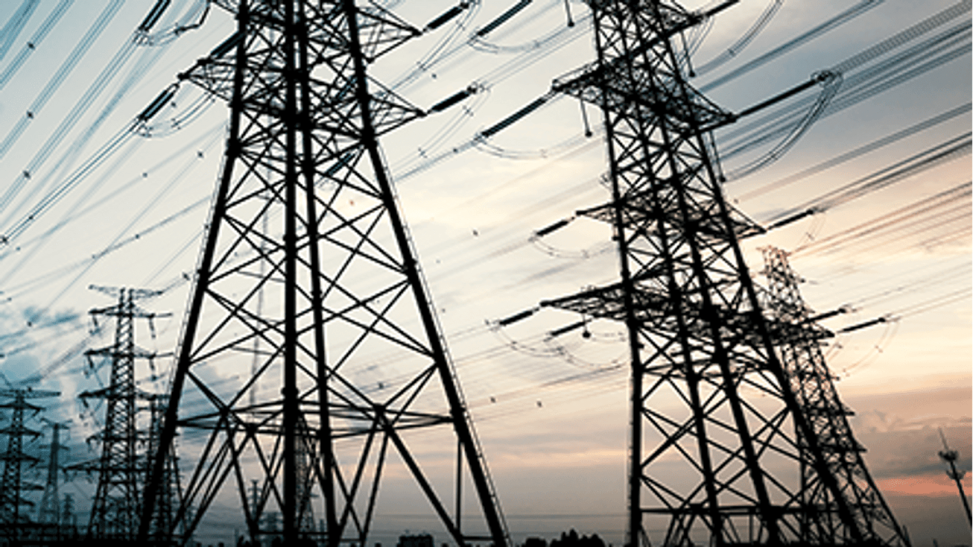 Multiple tall electricity pylons with numerous power lines stretch across the sky at sunset, silhouetted against a cloudy sky with fading light.