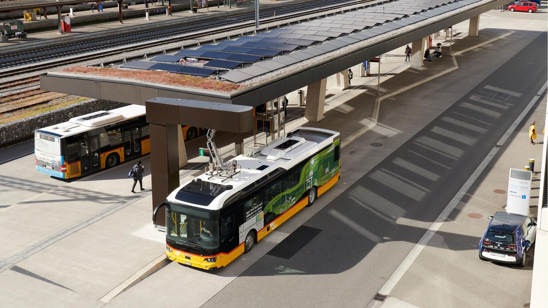 Ein gelber und grüner Elektrobus lädt an einer Busstation auf, angeschlossen an ein oberirdisches Ladegerät. Ein weiterer Bus und mehrere geparkte Autos sind in der Nähe, im Hintergrund sind die Bahngleise zu sehen.