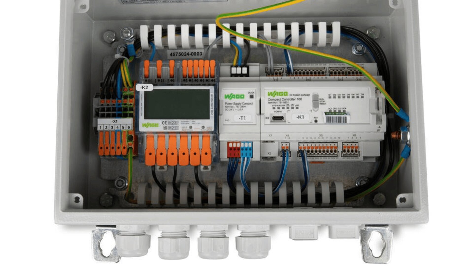 Ein elektrischer Schaltkasten mit verschiedenen beschrifteten Klemmen, Steckern, Drähten und elektronischen Modulen, einschließlich orangefarbener, blauer und grüner Komponenten, die übersichtlich angeordnet sind, um die Verkabelung und den Anschluss zu organisieren.