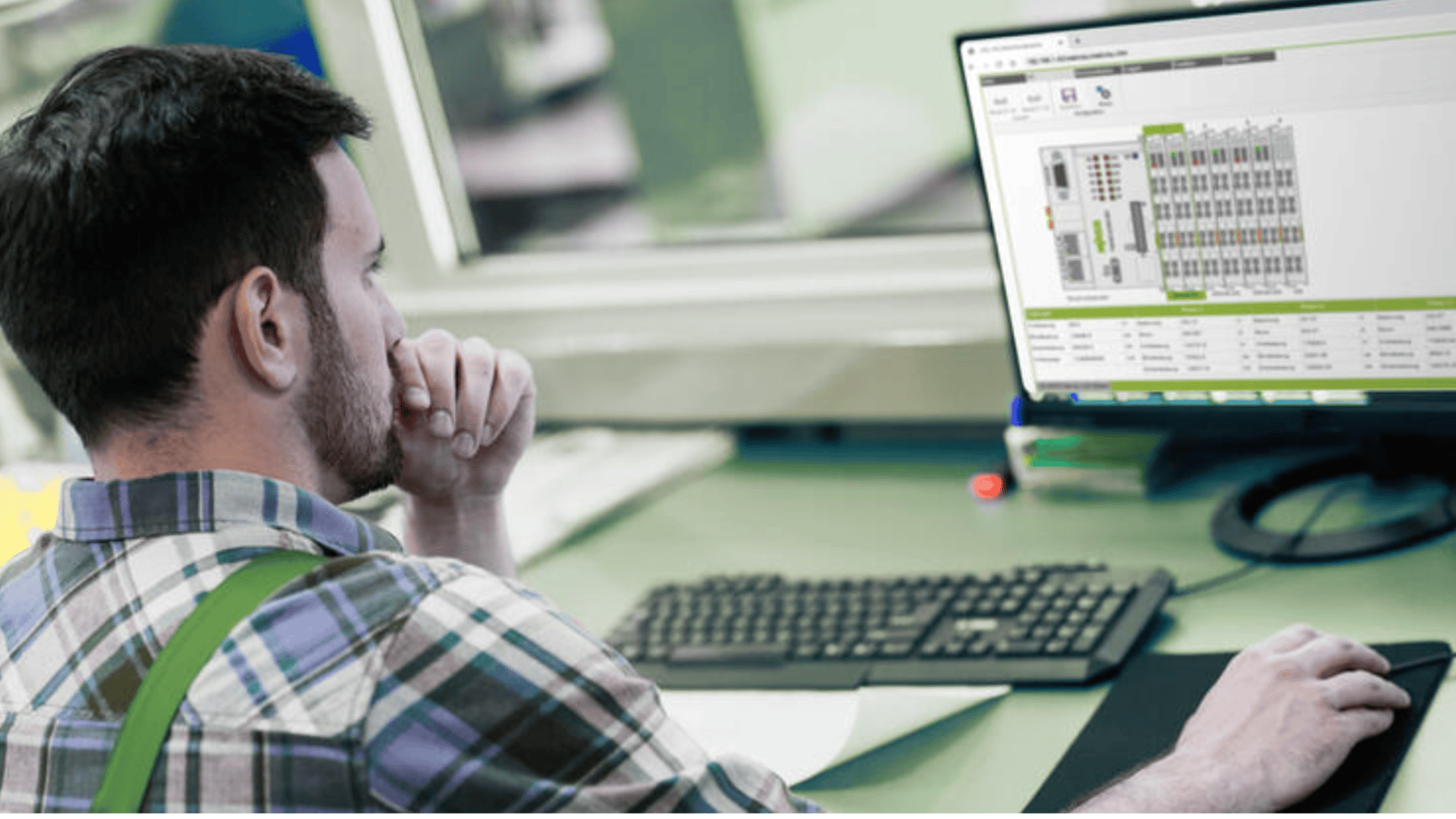 Ein Mann im karierten Hemd sitzt an einem Schreibtisch und schaut nachdenklich auf einen Computermonitor, auf dem technische Diagramme und Daten angezeigt werden. Vor ihm befinden sich eine Tastatur und eine Maus.