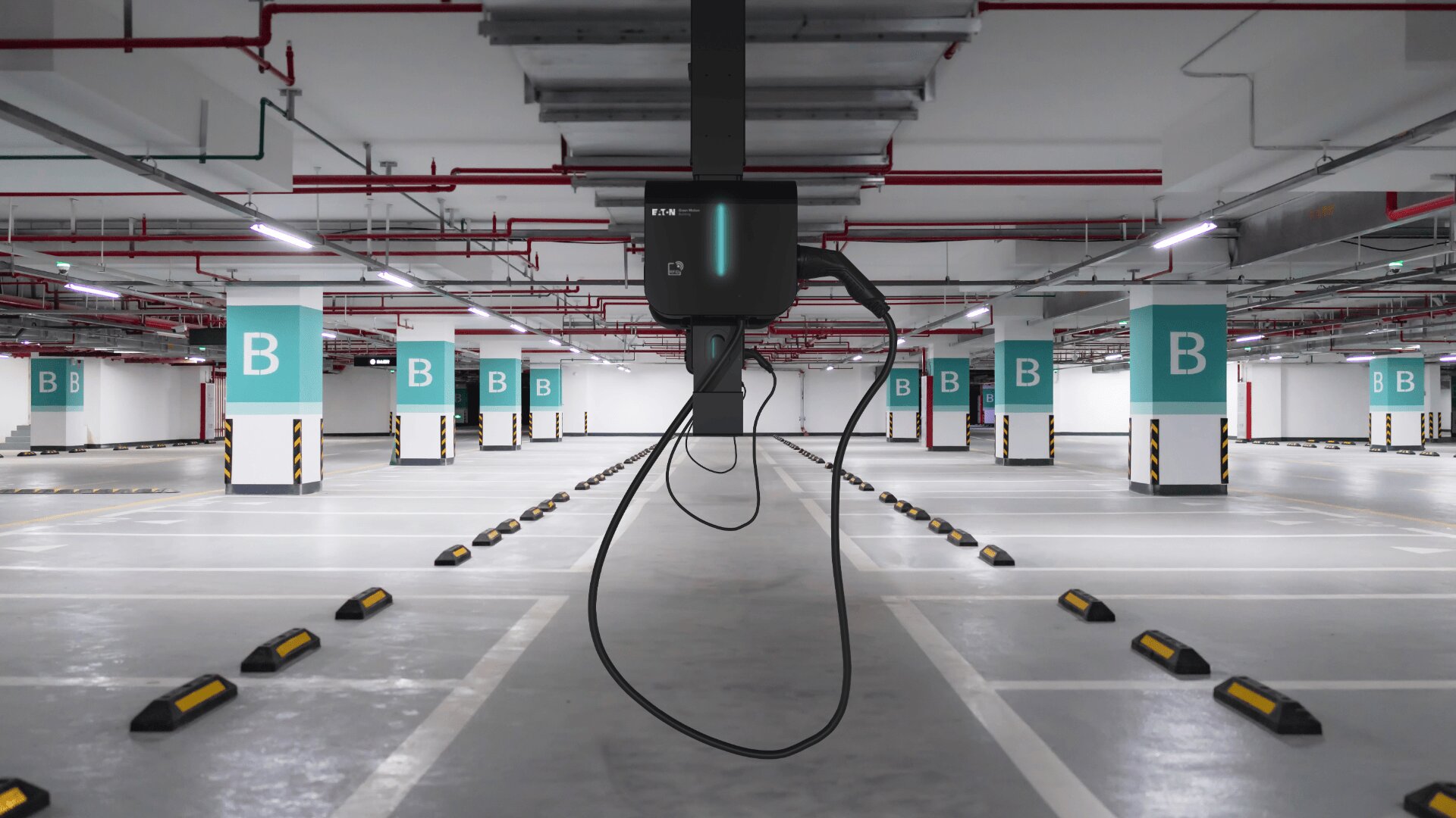 Eine leere Parkgarage mit weißen und blaugrünen Säulen mit der Aufschrift "B". In der Mitte des Vordergrunds hängt eine Ladestation für Elektrofahrzeuge mit Kabeln von der Decke. Schwarze und gelbe Parkbremsen säumen jeden Parkplatz.