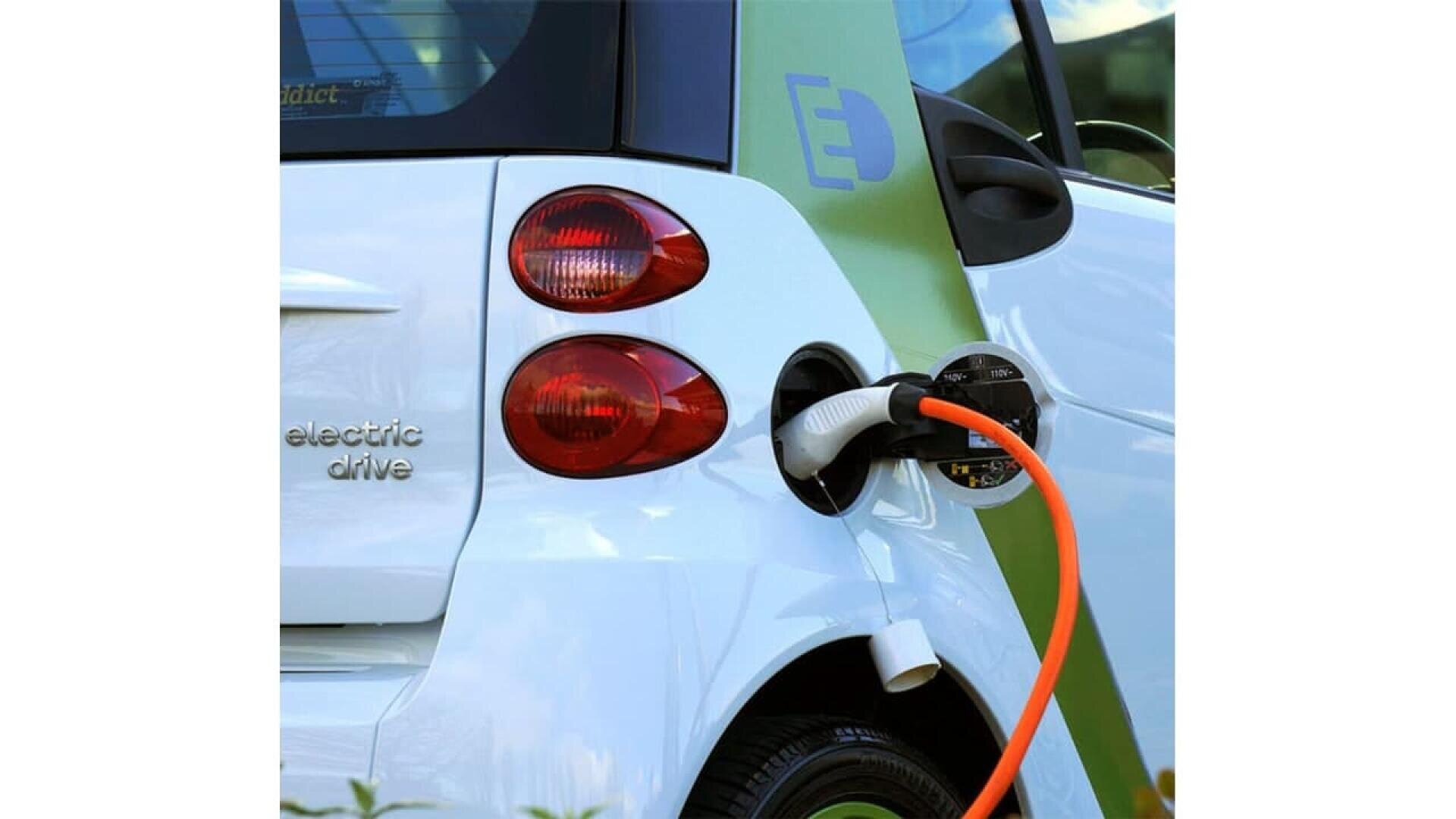 A close-up of a compact white electric car being charged, with an orange charging cable plugged into the side port near the taillights. The words electric drive are visible on the car.