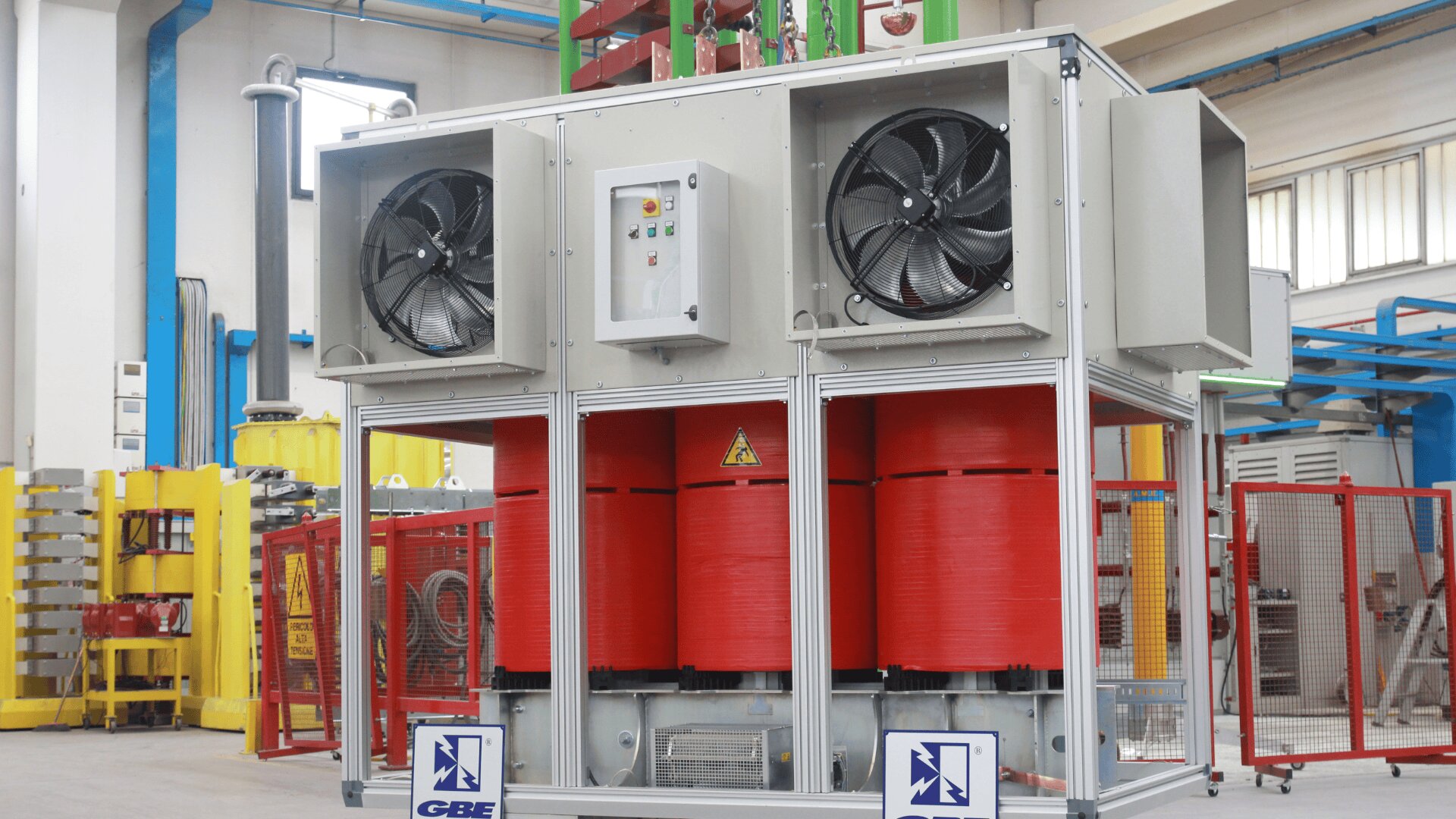 A large industrial transformer with red coils, protective casing, cooling fans, and control panel is situated in a factory setting with blue beams and other machinery visible in the background.