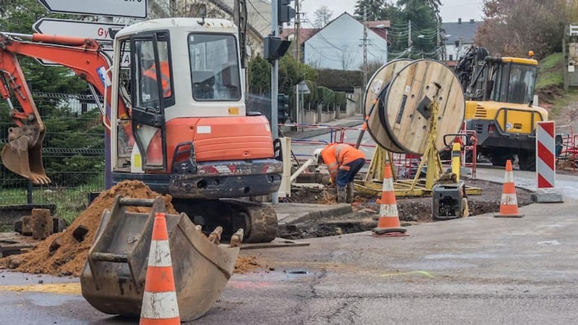 Two construction workers are working on a street excavation site, surrounded by orange cones, construction vehicles, a large cable spool, and piles of dirt. Residential houses are visible in the background.