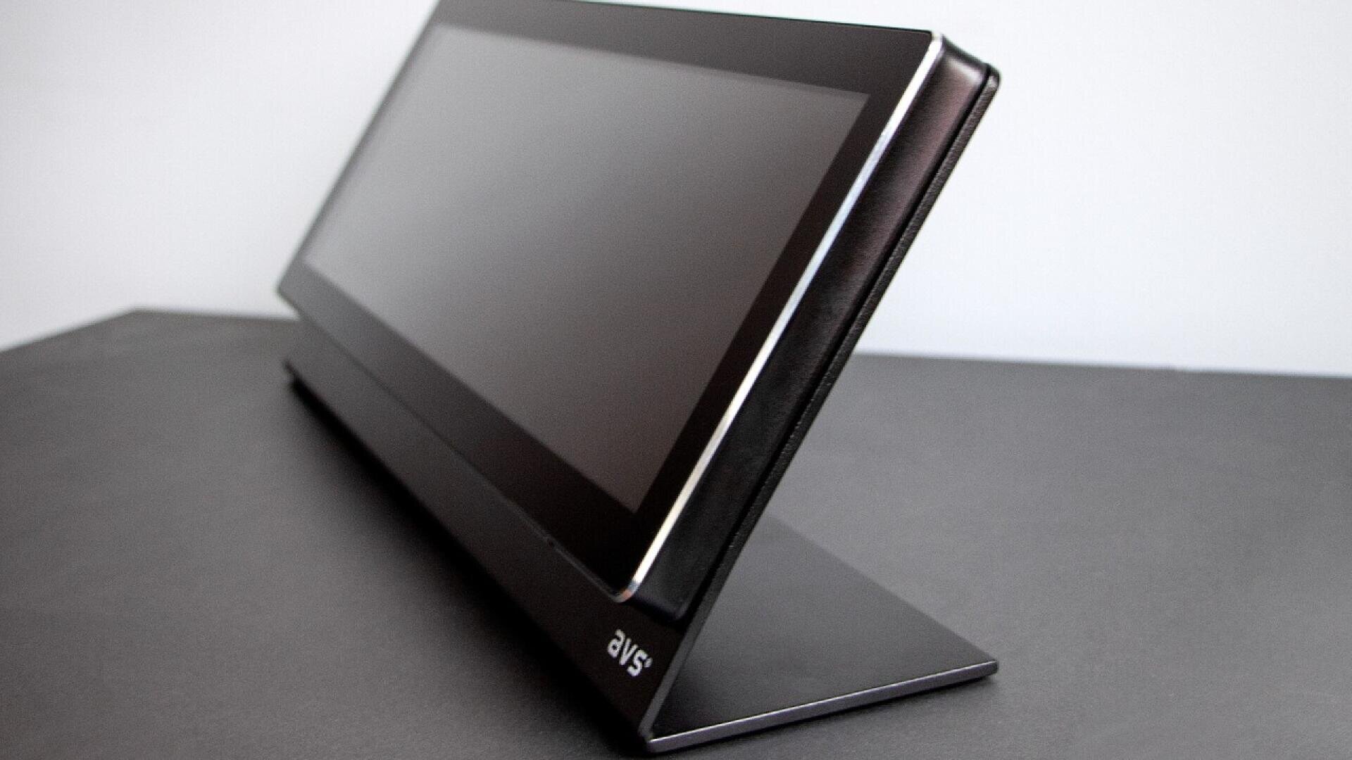 A thin, black portable monitor with a rectangular screen sits on a matching stand against a plain, light-colored background. The devices brand logo is visible on the bottom left corner of the stand.