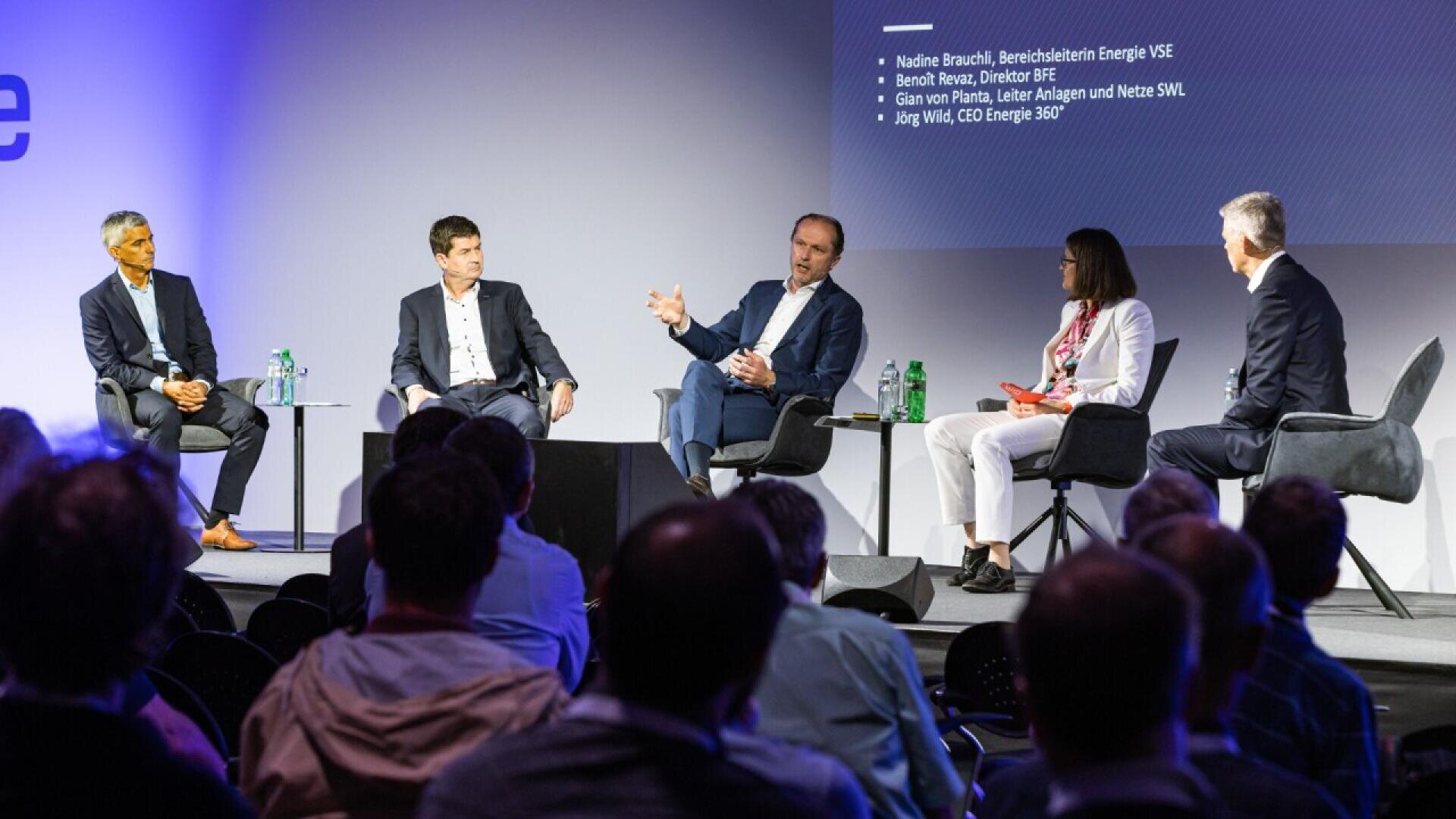 Five people sit on stage in a panel discussion, facing an audience. One man is speaking and gesturing with his hands, while the others listen. A screen displays names and titles in the background.