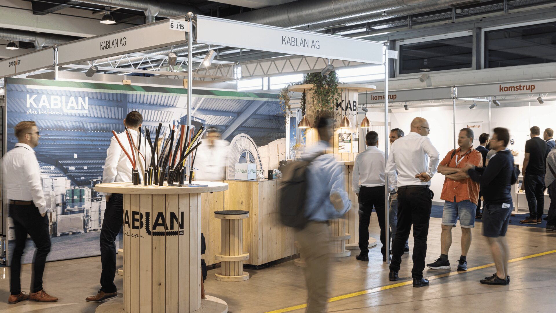 Eine geschäftige Messe mit Menschen, die am Stand der KABLAN AG interagieren, der aus einem hölzernen Display mit Kabeln und einer großen Kulisse besteht. Andere Messestände sind im Hintergrund zu sehen.
