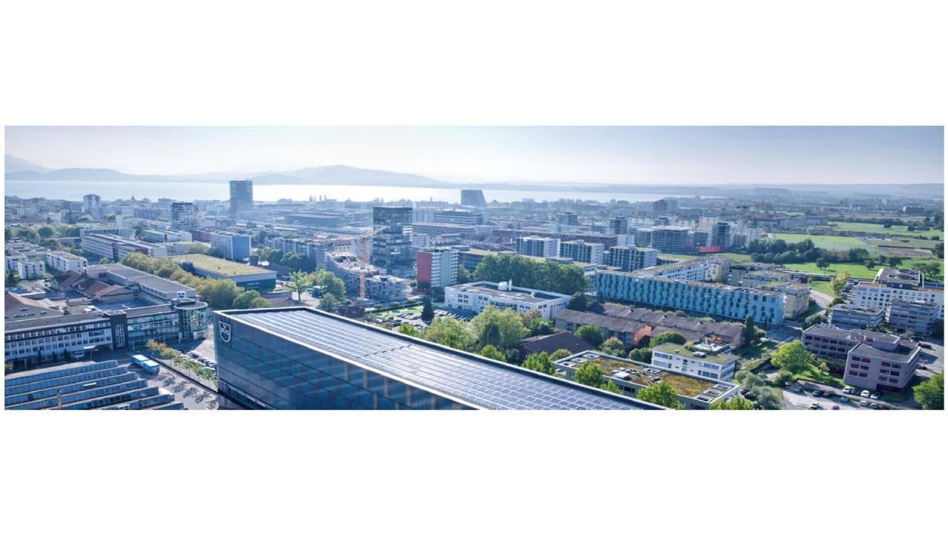 A wide aerial view of a modern city with many buildings, green areas, and distant mountains near a large body of water under a clear blue sky.