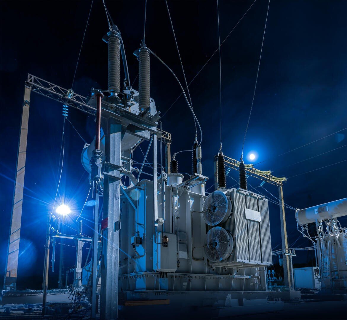 High-voltage electrical substation at night, illuminated by blue lighting, with large transformers, insulators, and power lines set against a dark sky with the moon visible in the background.