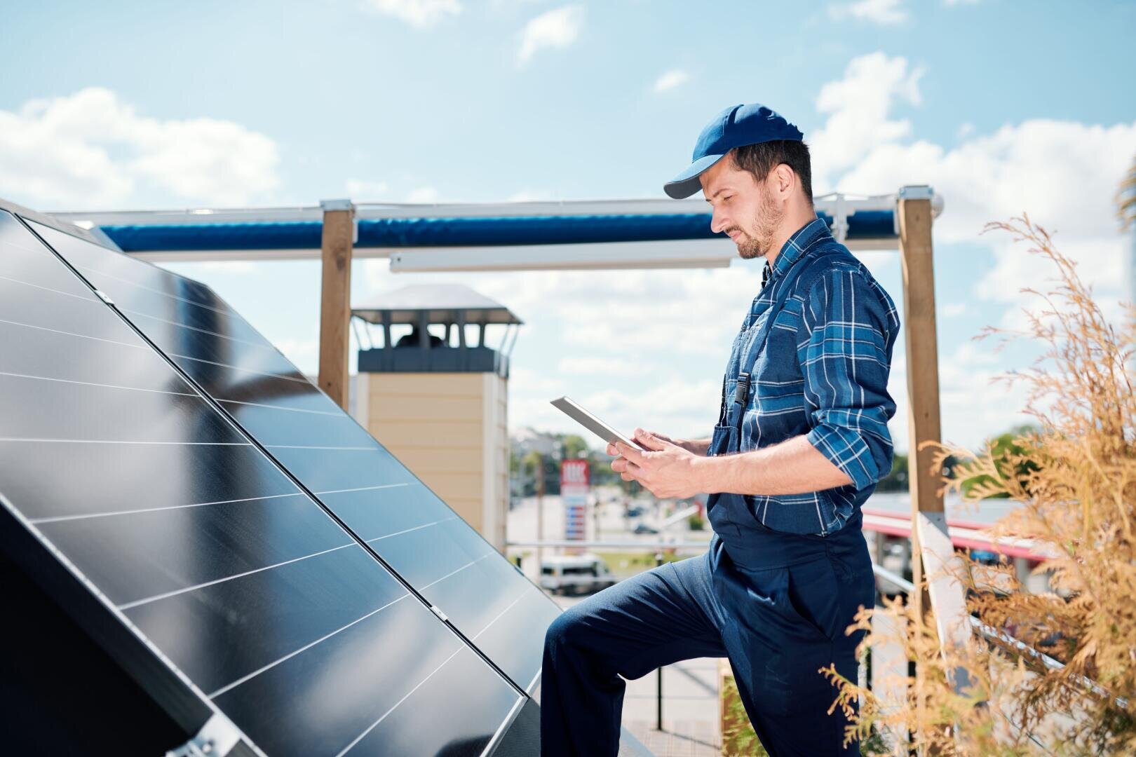 Ein Mann in Arbeitskleidung und Mütze inspiziert an einem sonnigen Tag große Solarpaneele im Freien, hält ein Tablet in der Hand und macht sich Notizen; im Hintergrund sind Pflanzen und Gebäude zu sehen.
