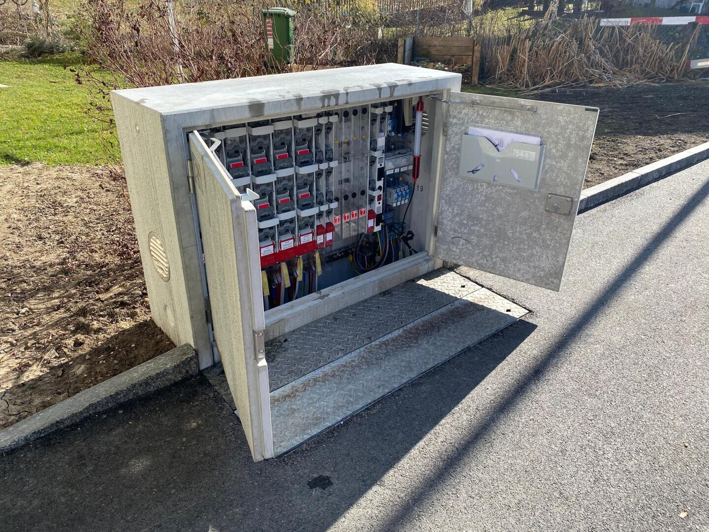 Ein grauer Stromkasten mit geöffneter Tür, die den Blick auf elektrische Schalttafeln, Schalter und Kabel im Inneren freigibt. Der Kasten ist auf einem Bordstein neben einer Straße installiert, im Hintergrund sind Gras und Erde zu sehen.