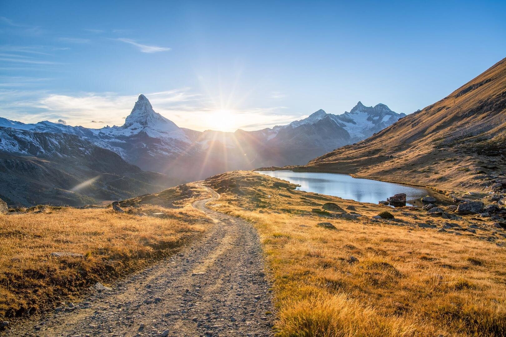 Ein unbefestigter Weg führt zu einem kleinen See und schneebedeckten Bergen bei Sonnenaufgang. Goldenes Gras bedeckt die Landschaft und die Sonne scheint knapp über dem Horizont.