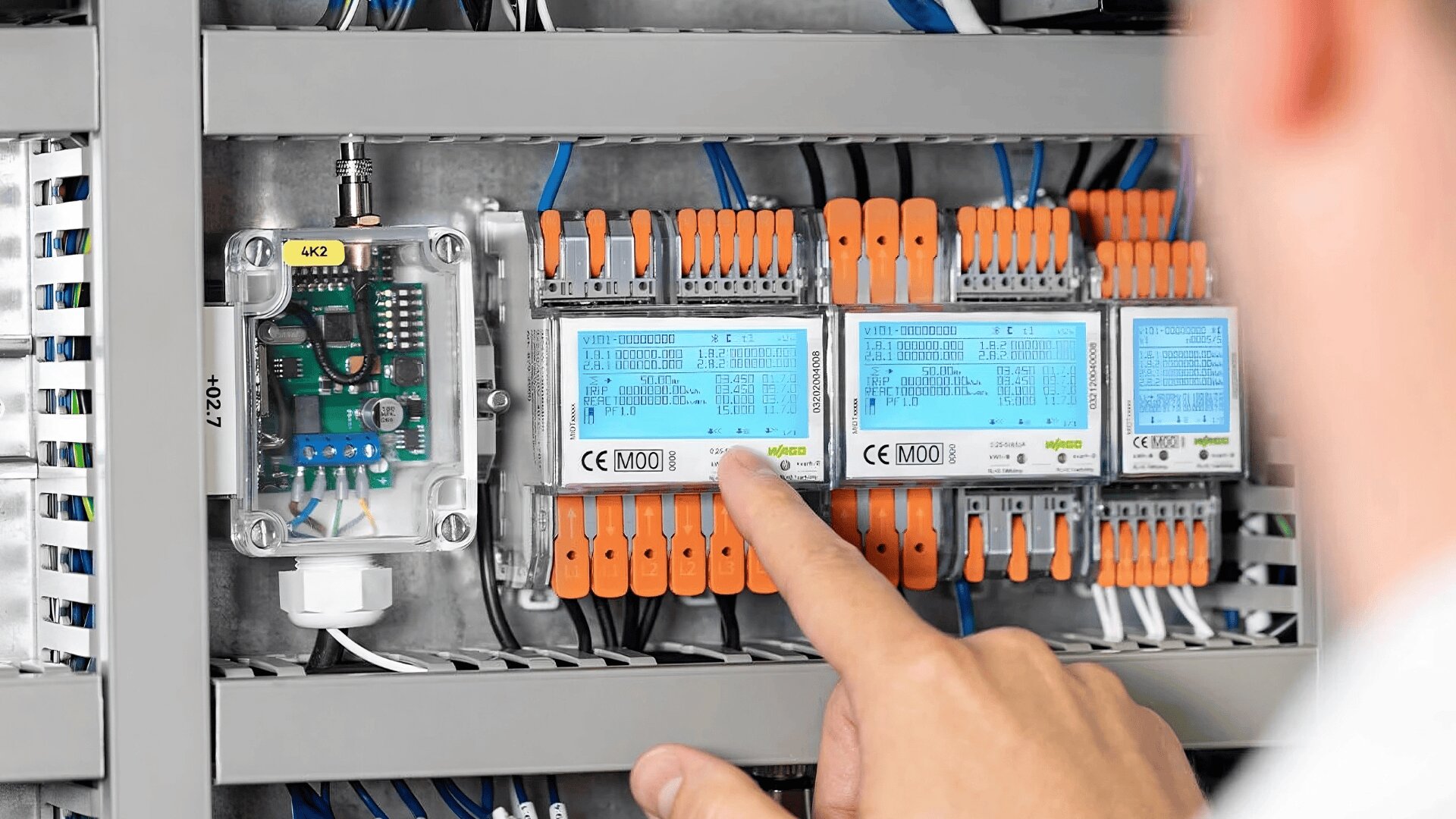 Die Hand einer Person zeigt auf den Bildschirm eines elektrischen Schaltschranks mit einer Vielzahl von Drähten, Klemmen und Bauteilen, die ordentlich in einem Metallgehäuse angeordnet sind.